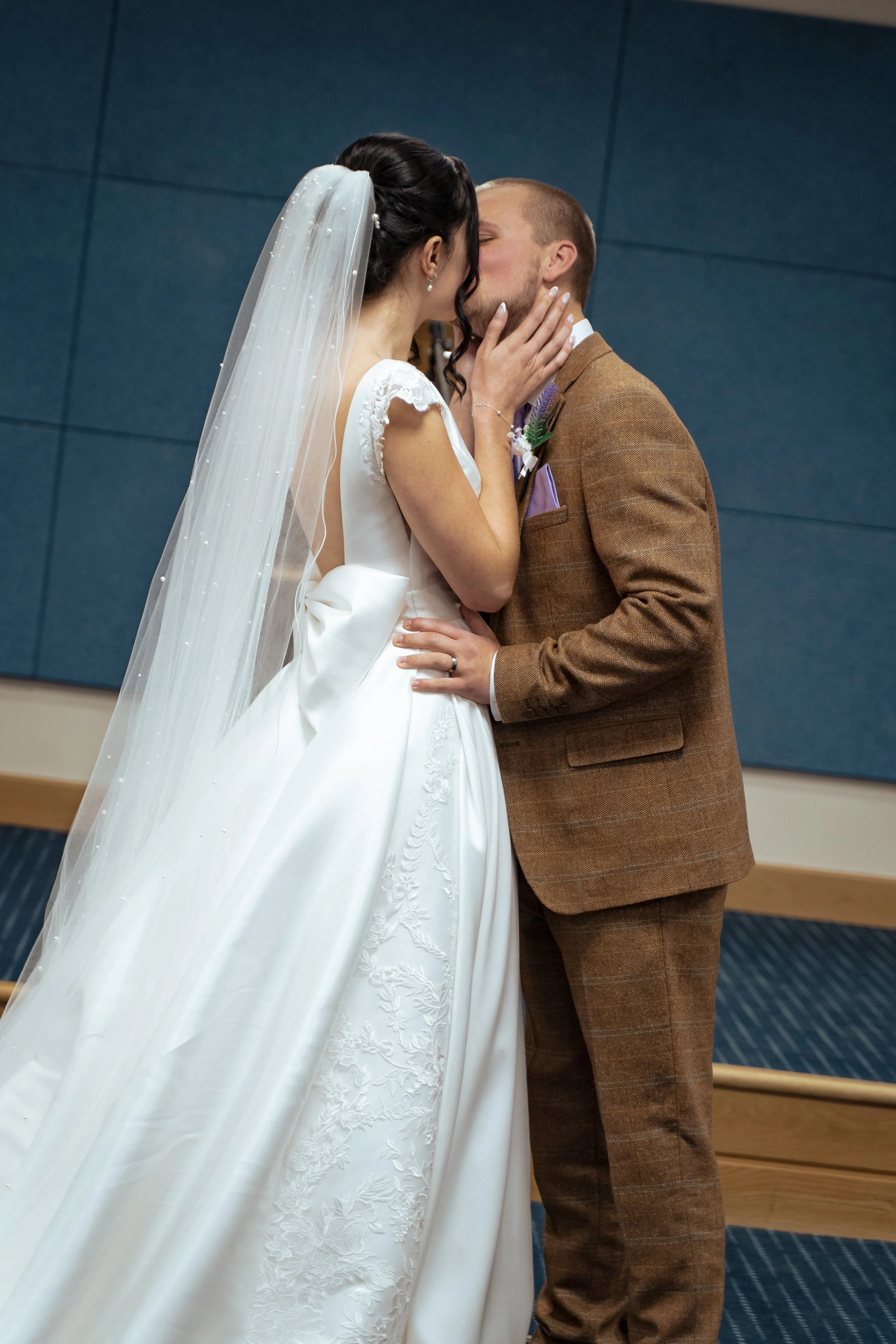 A bride and groom sharing a kiss during their wedding ceremony indoors. The bride is wearing a white wedding dress with lace details and a veil, while the groom is dressed in a brown suit with a purple tie and boutonniere.