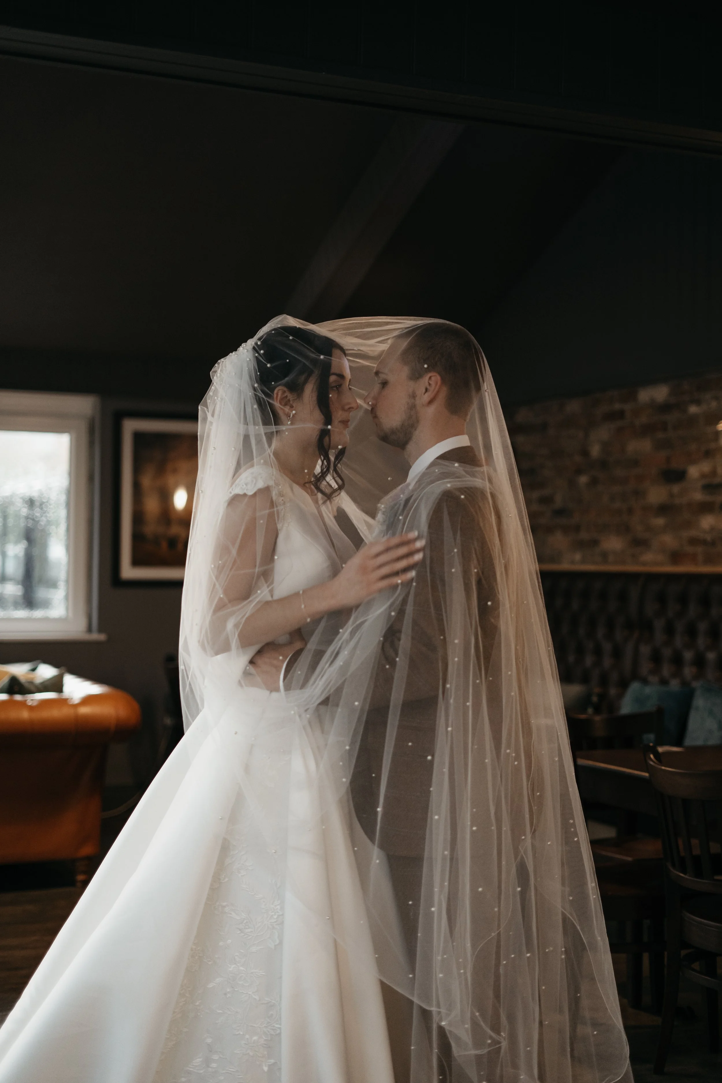Bride and groom standing face to face behind a wedding veil, inside a room with brick wall and window in the background.