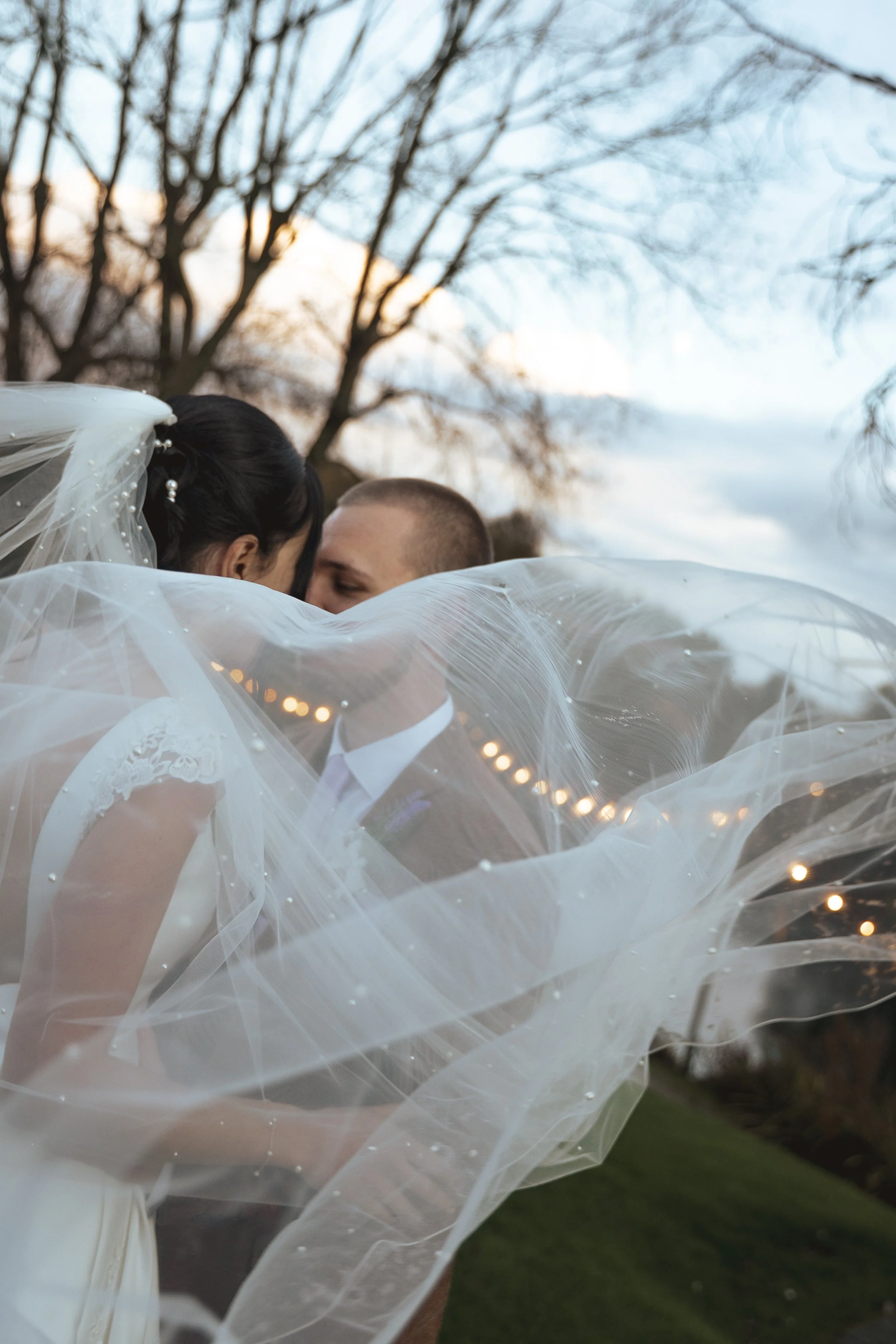 A bride and groom kissing outdoors, with the bride wearing a veil with pearls and lace, and the groom in a suit, surrounded by flowing white fabric and fairy lights.