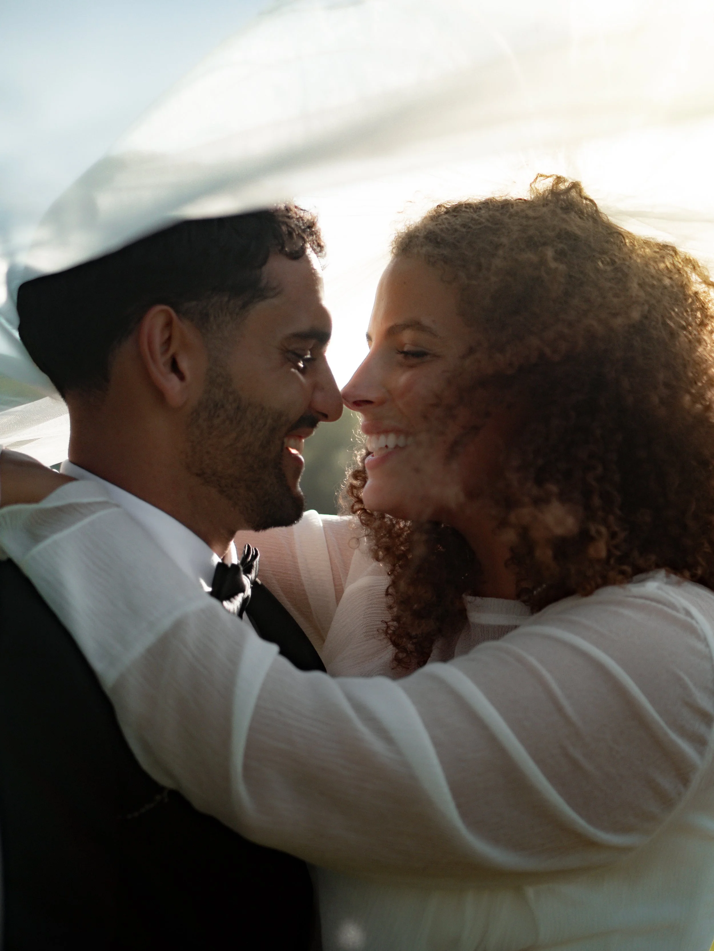 A close-up photo of a smiling couple with their foreheads touching, dressed in formal attire, outside with sunlight illuminating their faces.