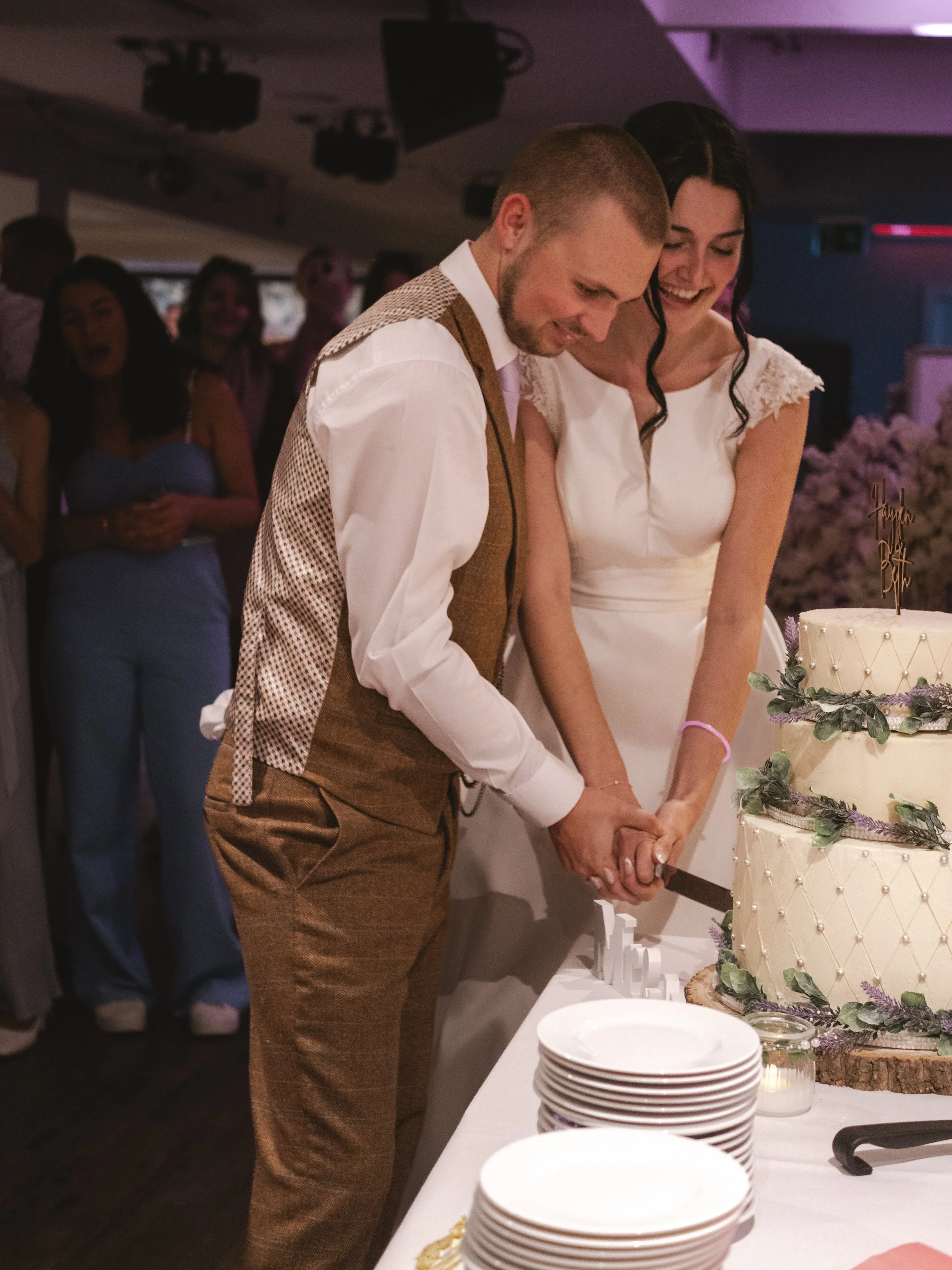 A man and a woman cutting a wedding cake together at their wedding reception, with guests in the background observing.