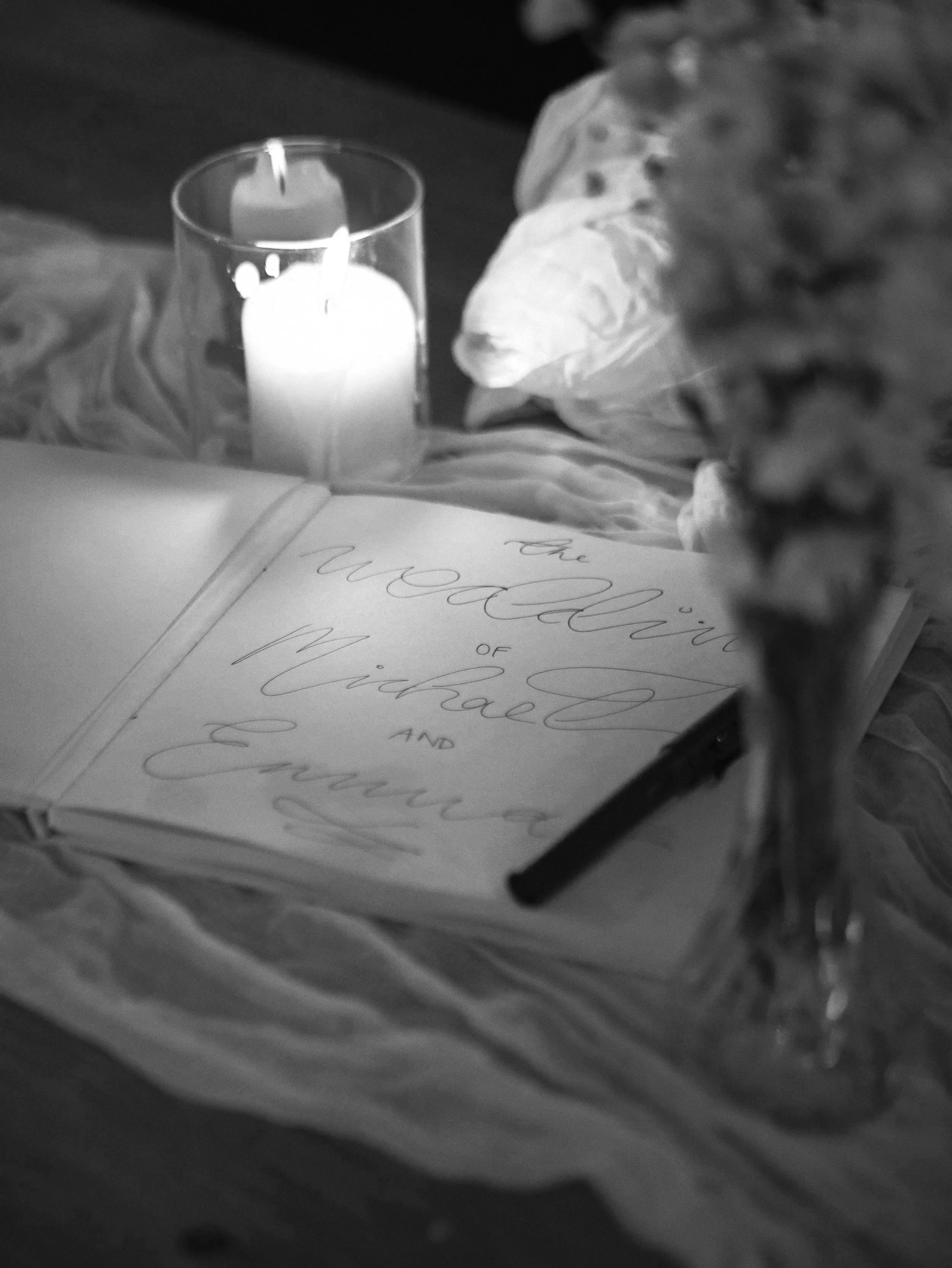 A lit white candle in a glass holder on an open book with cursive writing, surrounded by flowers.