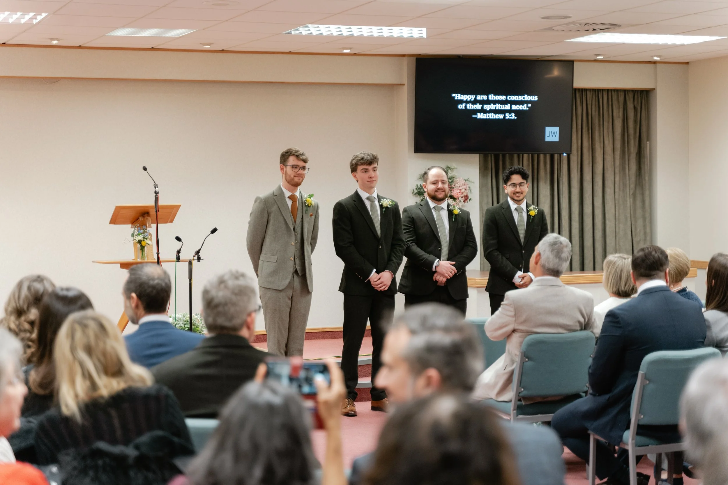 Four young men dressed in suits and wearing boutonnieres standing at the front of a room with an audience, likely at a wedding or formal event. A large screen displays a Bible quote from Matthew 5:3, and a podium decorated with flowers is visible to 