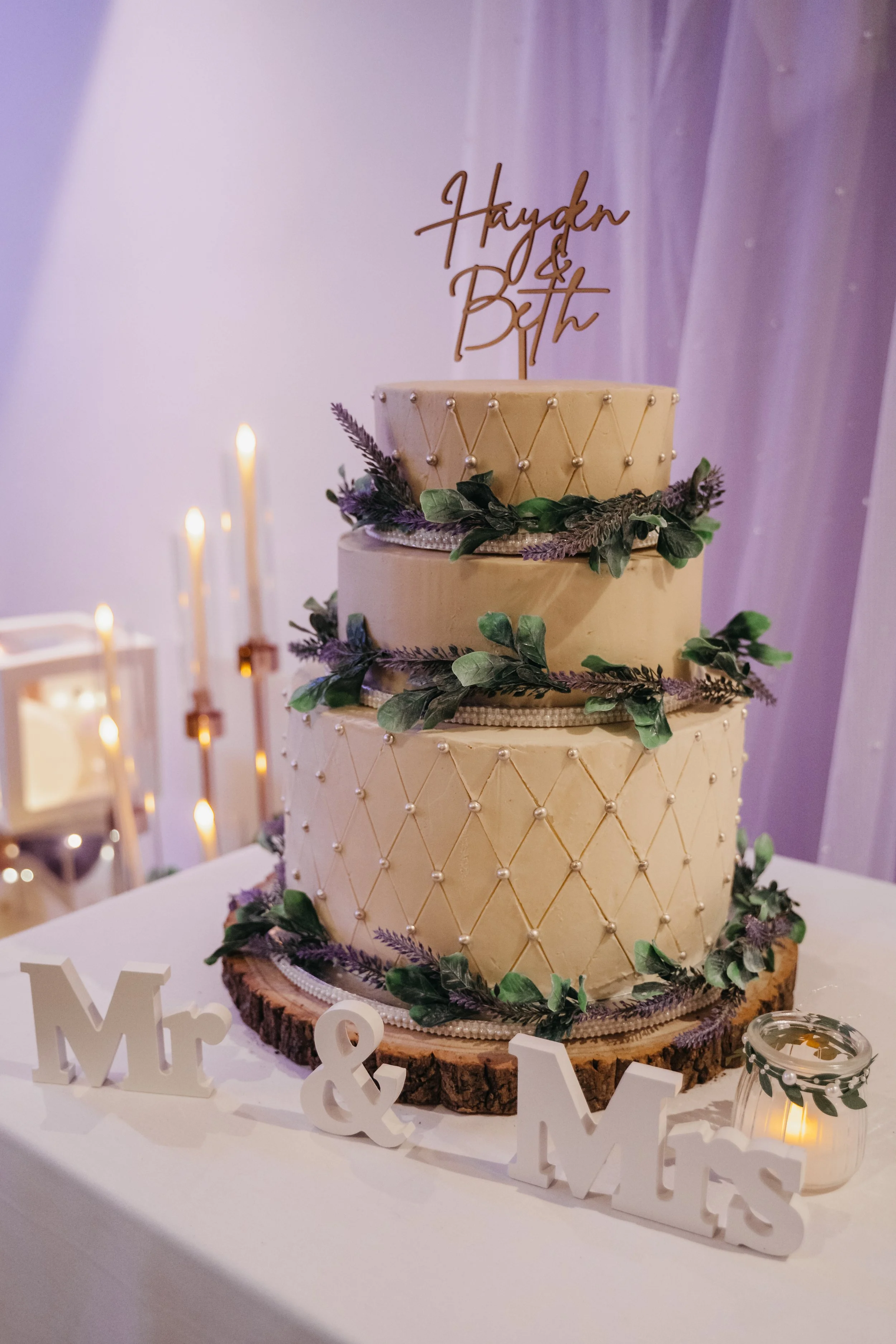 A three-tier wedding cake decorated with purple flowers and green leaves, with a gold cake topper reading "Häyden & Birt." The cake sits on a wooden base with tree bark texture, surrounded by white tables, candles, and decorative signs reading "Mr & 