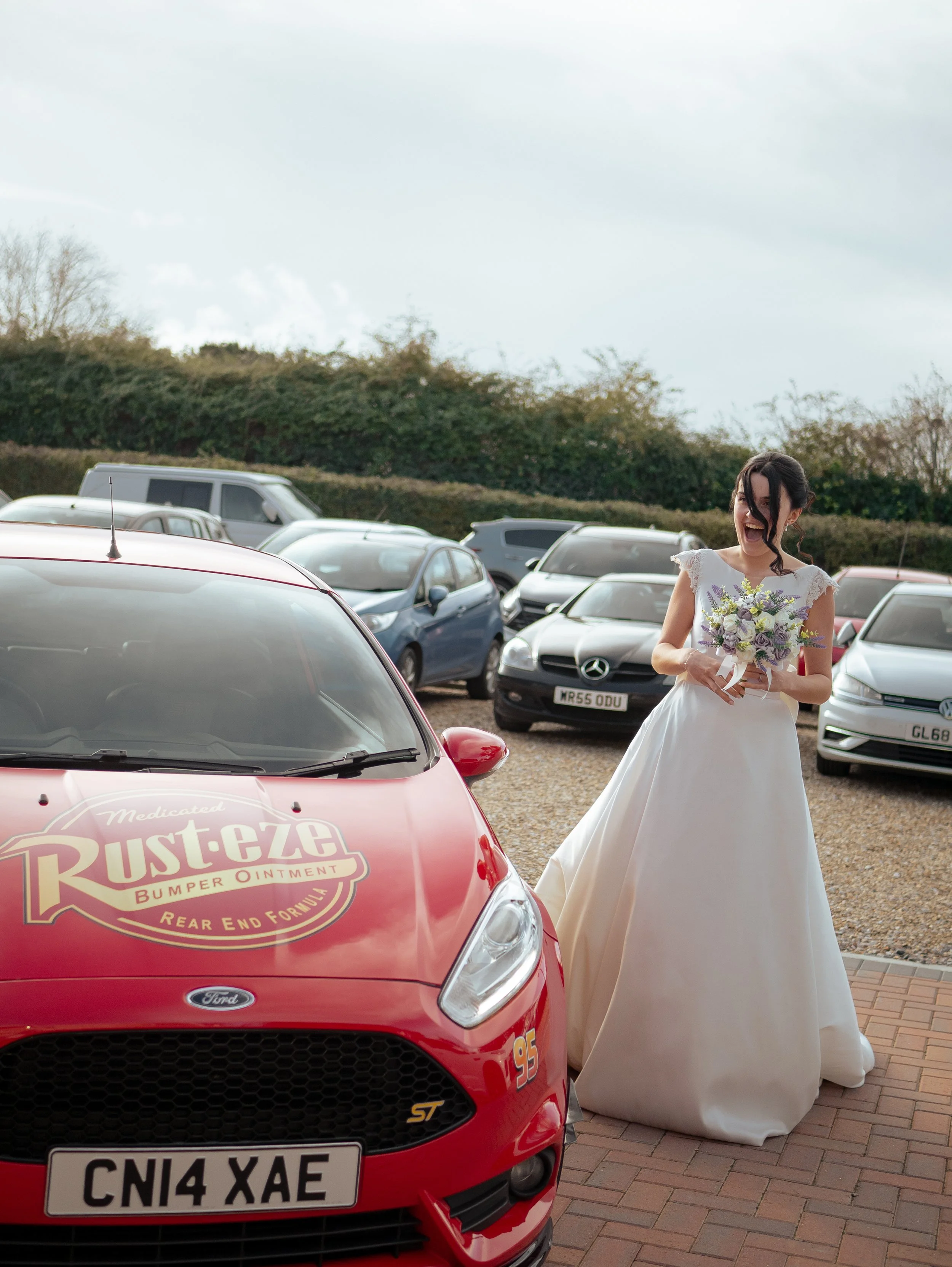 A woman in a white wedding dress holding a bouquet of flowers, standing next to a red race car in a parking lot, with multiple cars and a hedge in the background.
