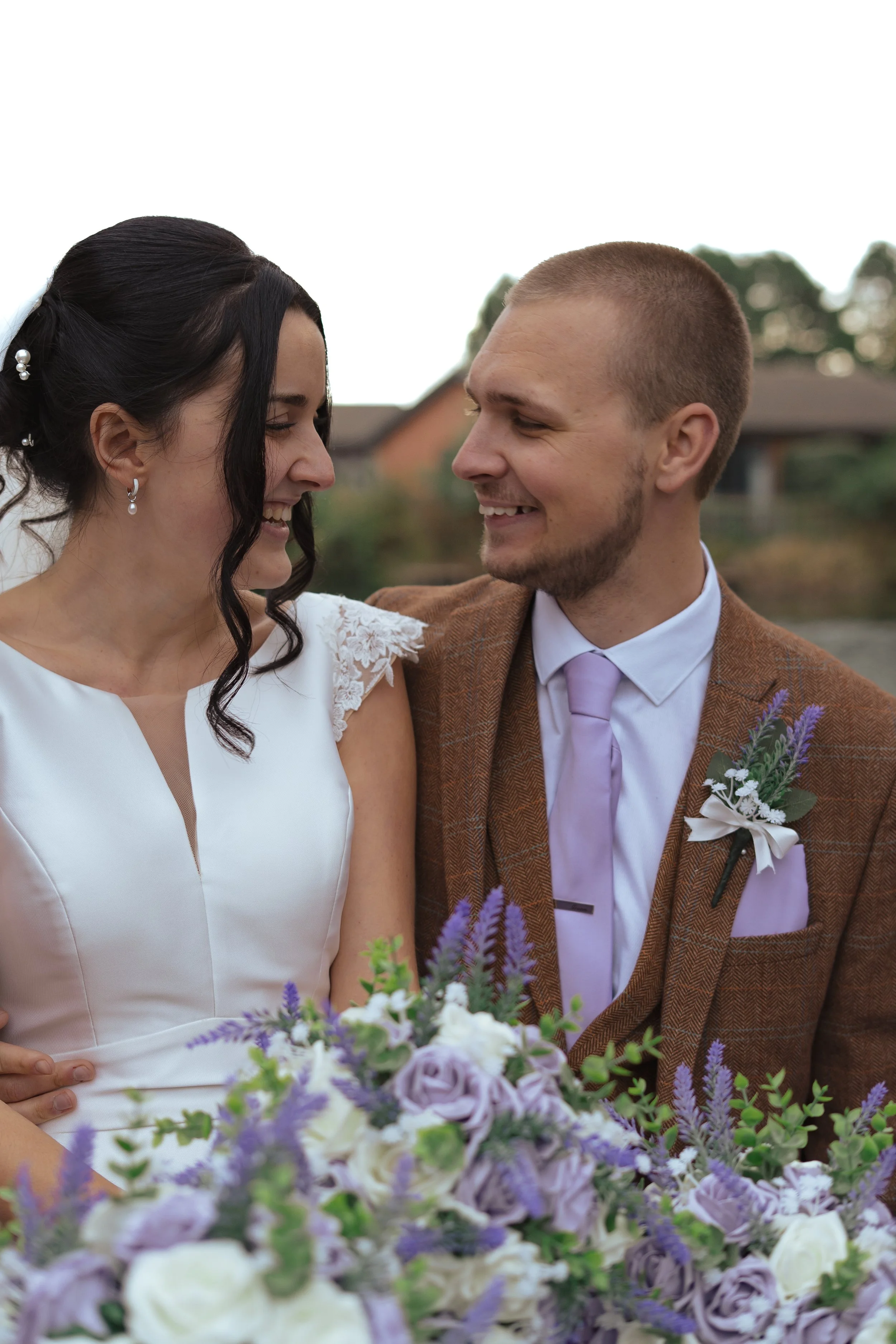 A happy couple looks at each other outdoors, with a wedding bouquet of purple and white flowers in the foreground.