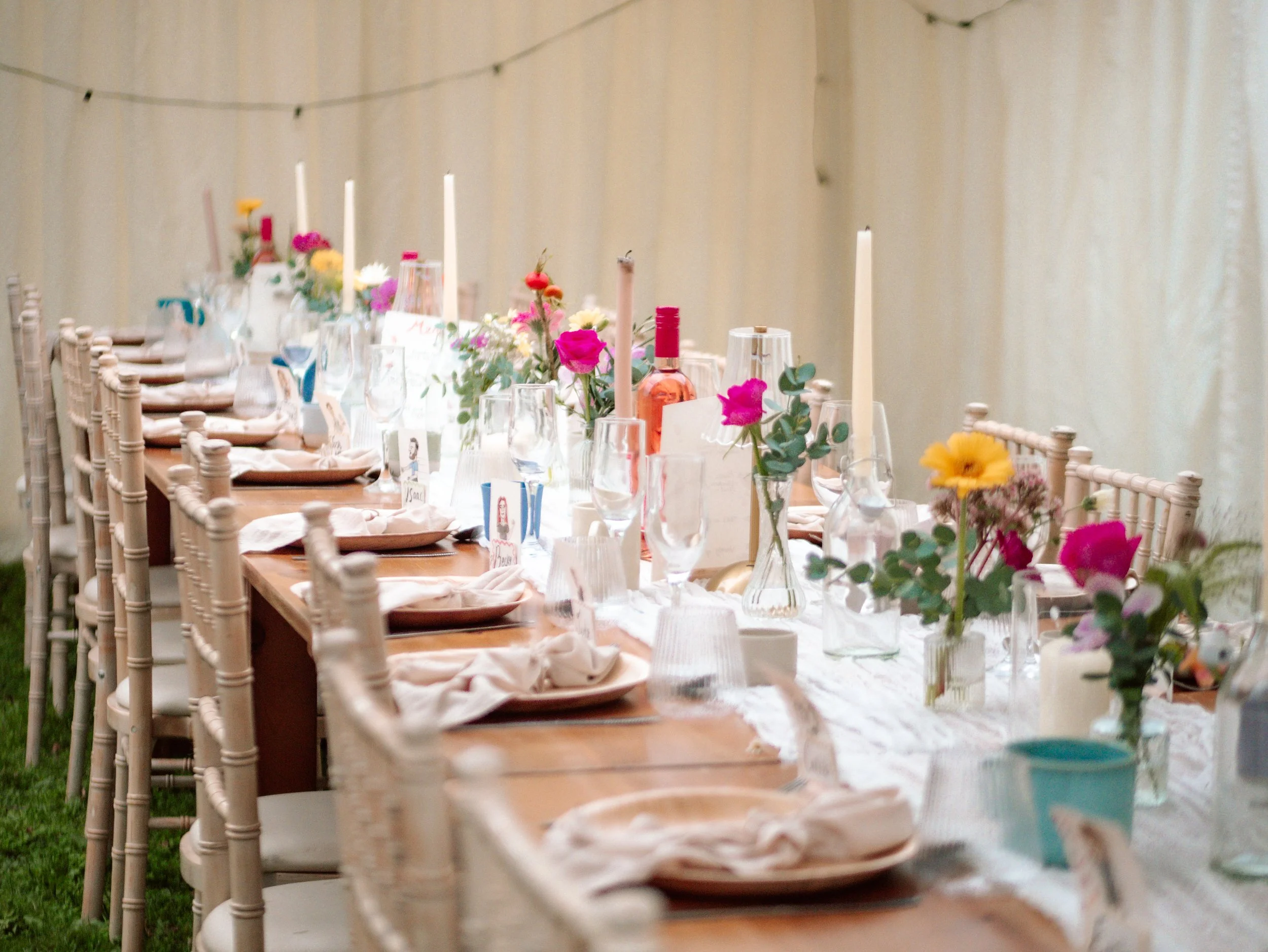 A long banquet table set up outdoors with chairs, floral centerpieces, candles, and place settings, prepared for a celebration or event.