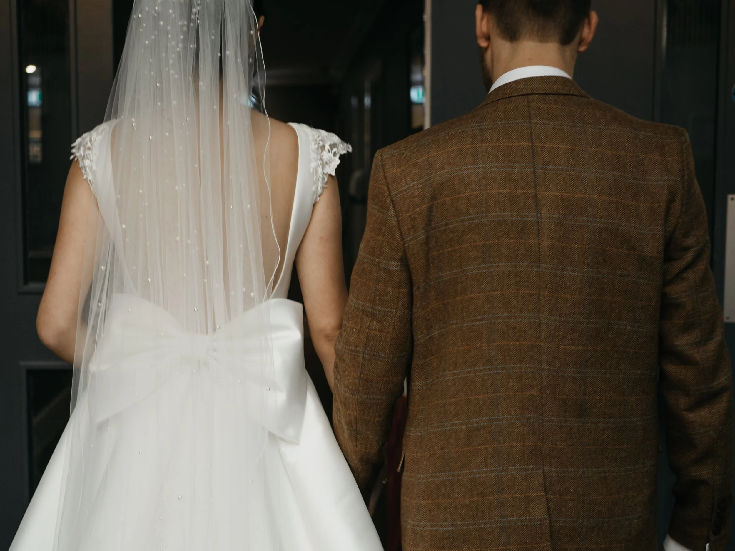 Back view of a bride and groom walking together, the bride wearing a white wedding dress with lace details and a veil, the groom wearing a brown checkered suit.