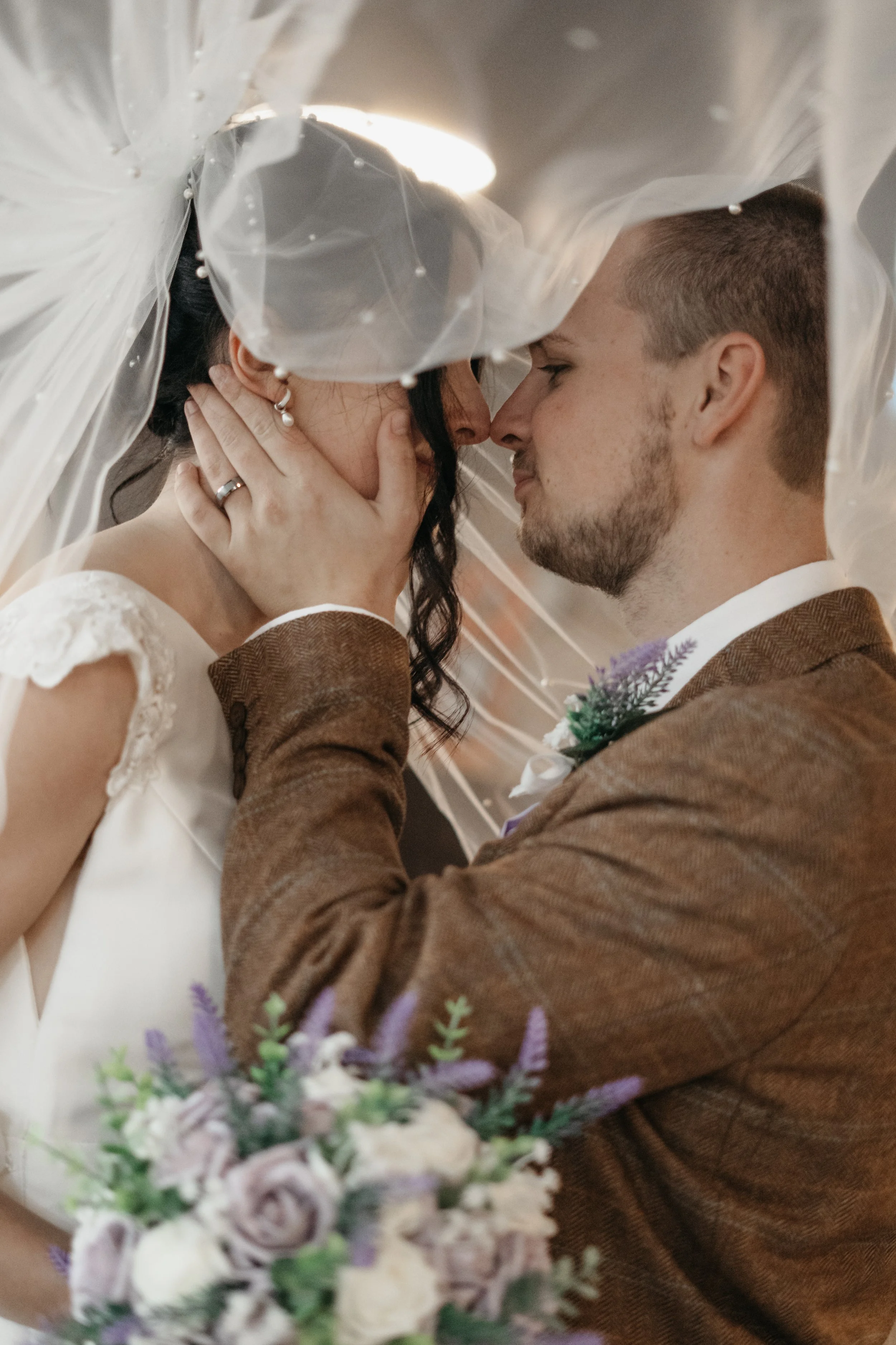 A man and woman are about to kiss beneath a wedding veil, with the woman holding a bouquet of flowers.
