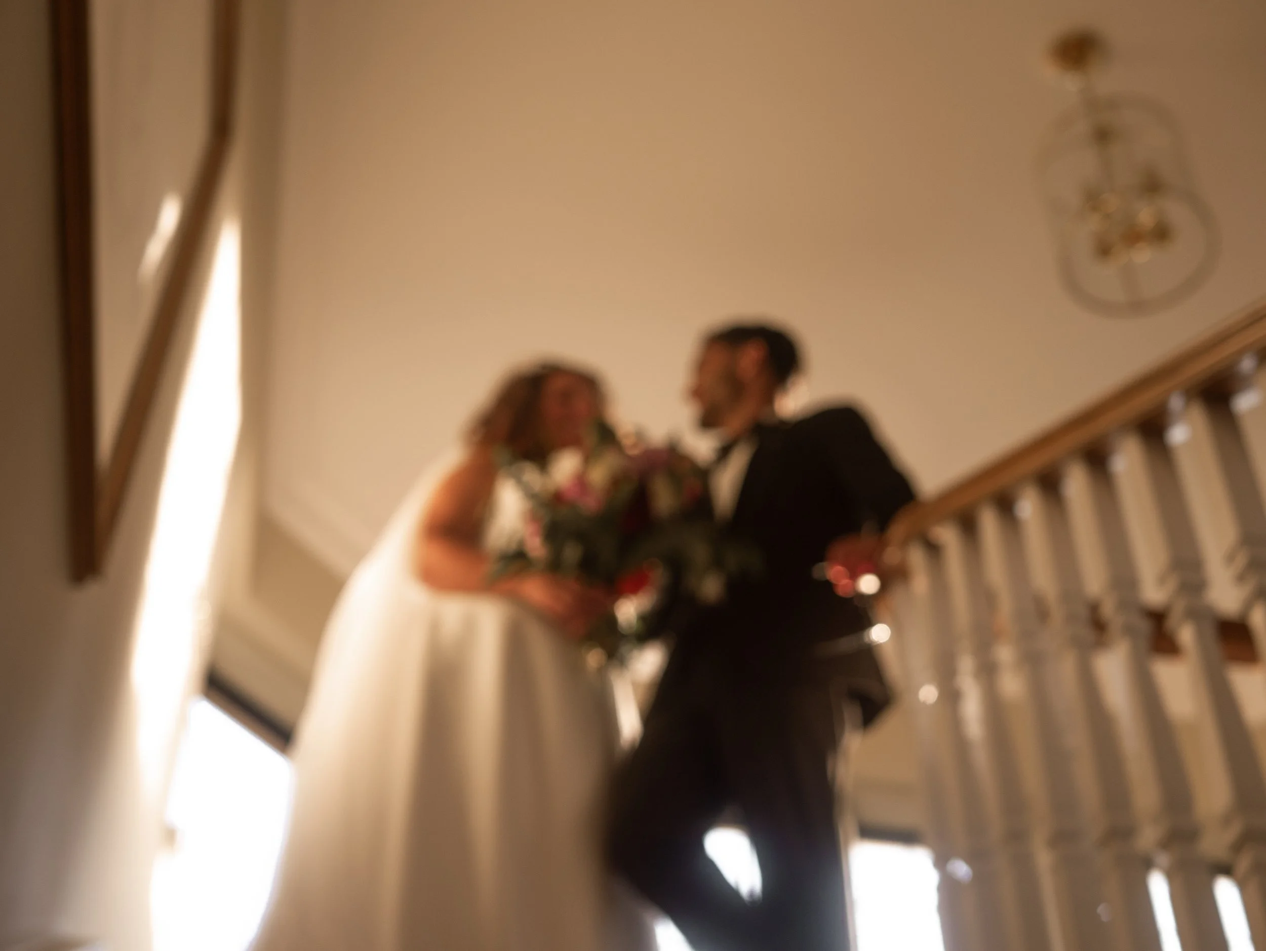Blurred photo of a couple standing close together on a staircase, with a woman holding a bouquet of flowers and a man wearing glasses, in an indoor setting with warm lighting.