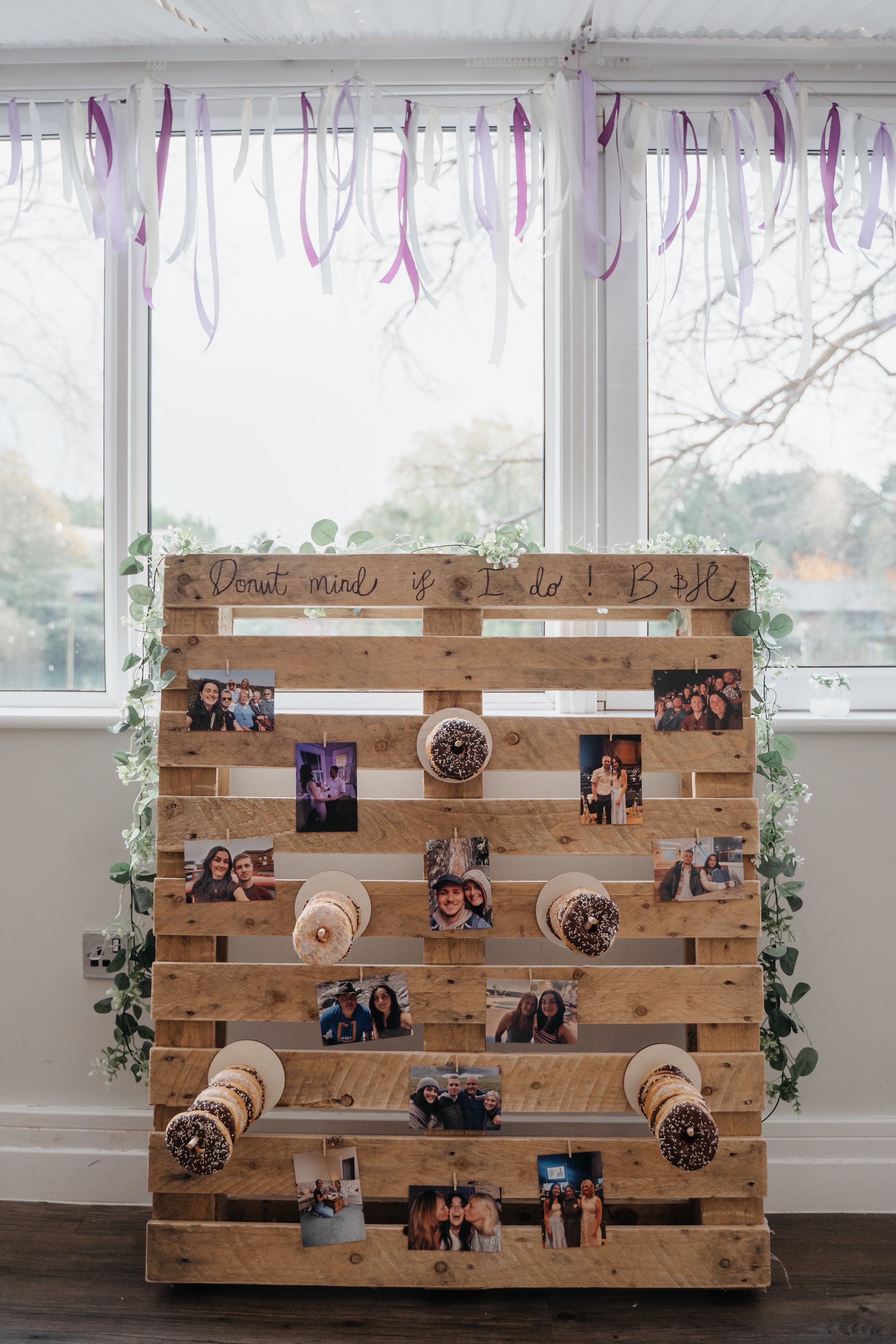 Wooden pallet with photos, donuts, and a handwritten sign that says 'Don't mind if I do! B+B' in front of a window with hanging purple and white ribbons, decorated with green foliage.