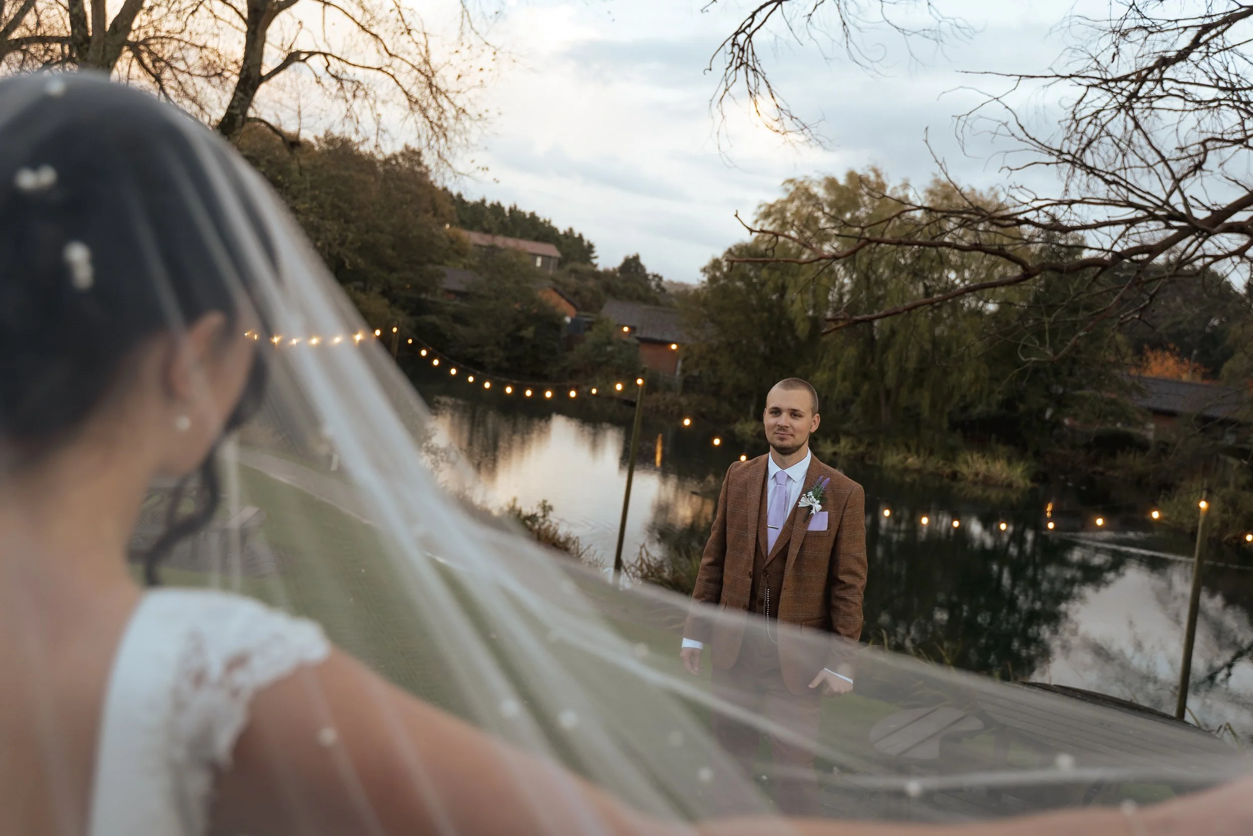 A groom standing by a river during sunset, well-dressed in a brown suit with a boutonniere, looking towards a bride who is capturing the moment with a wedding dress and veil, with trees and houses in the background, and string lights along the riverb