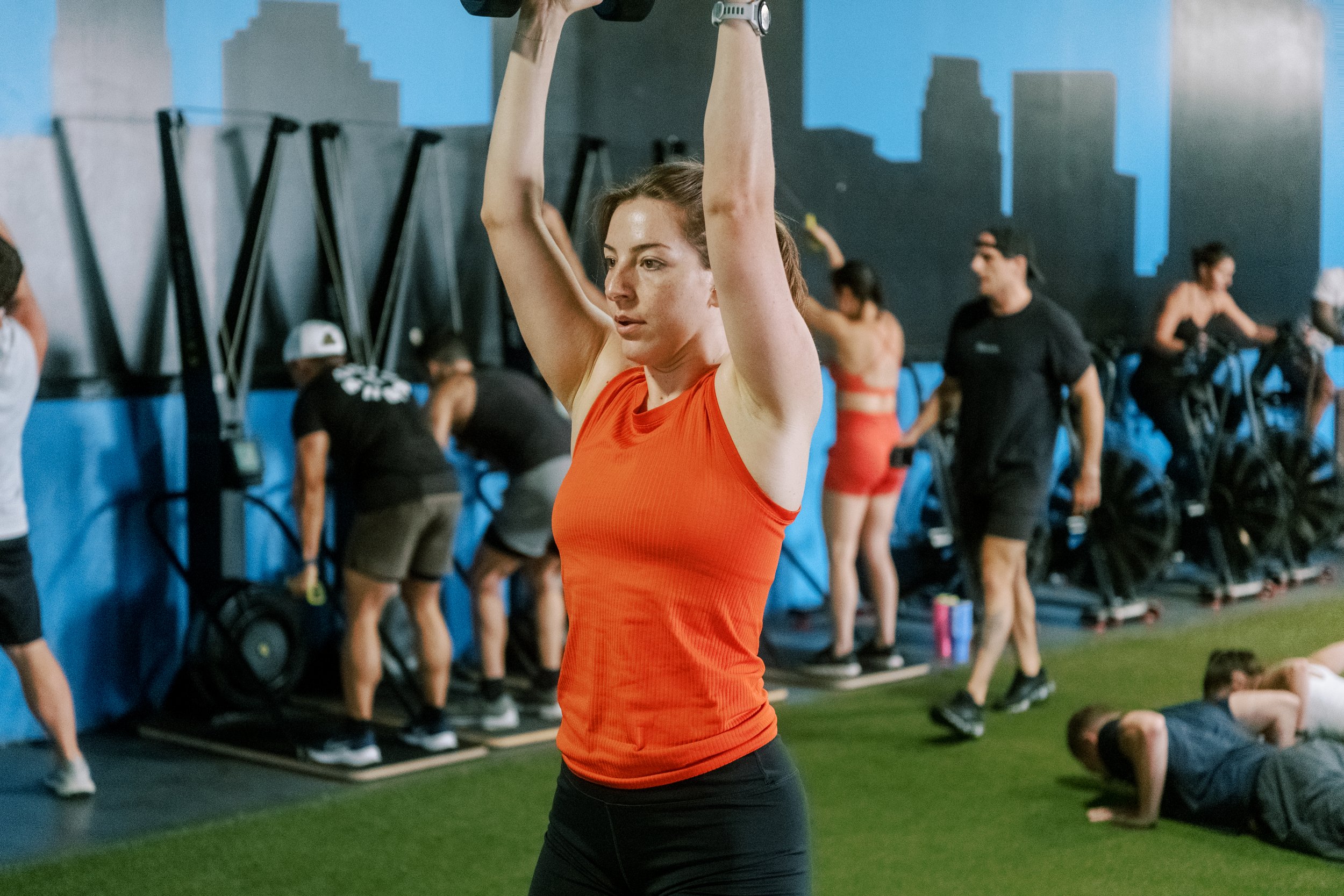 A woman with brown hair in a ponytail wearing an orange tank top and black leggings lifting a dumbbell overhead during a workout session at a gym. In the background, other individuals are engaging in various exercises, including lifting weights and doing push-ups, under a mural of a city skyline.