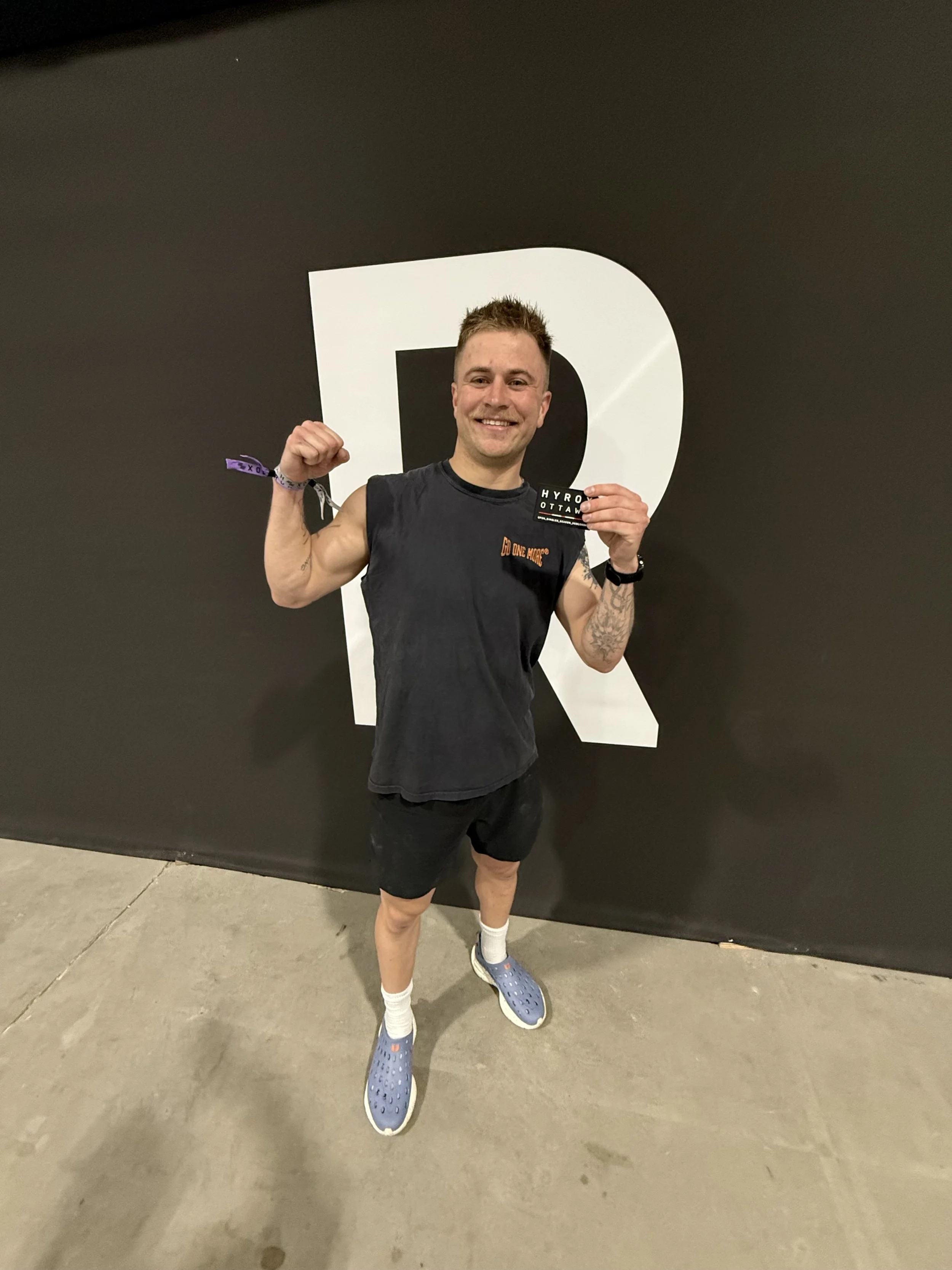 A young man dressed in athletic clothing poses confidently with a clenched fist, smiling at the camera, standing in front of a large letter R on a dark wall, wearing blue slip-on shoes, white socks, and a black wristwatch in a gym or sports facility.