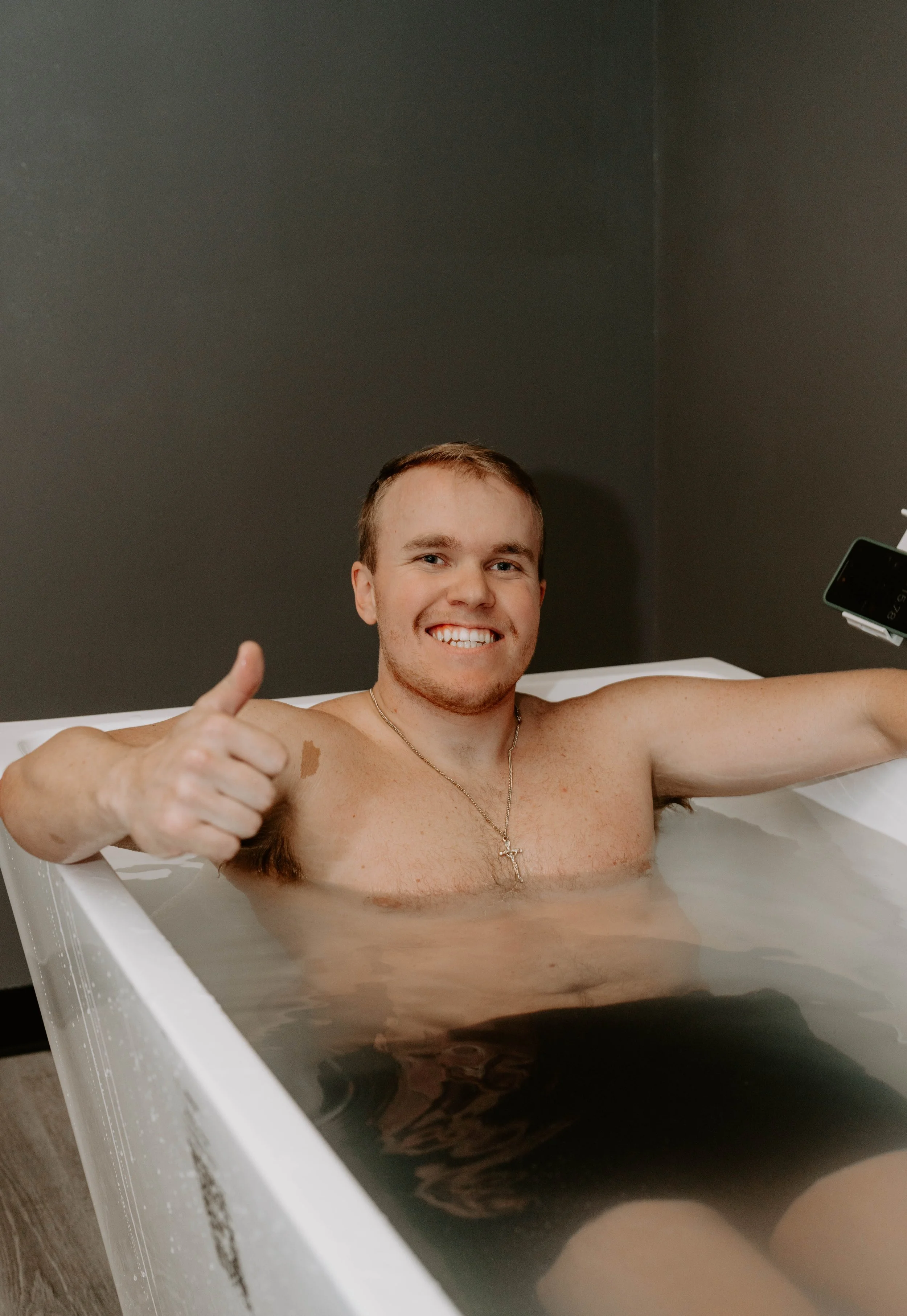 A smiling young man with short brown hair in a bathtub, giving a thumbs-up with his right hand, wearing a cross necklace, with a gray wall in the background.