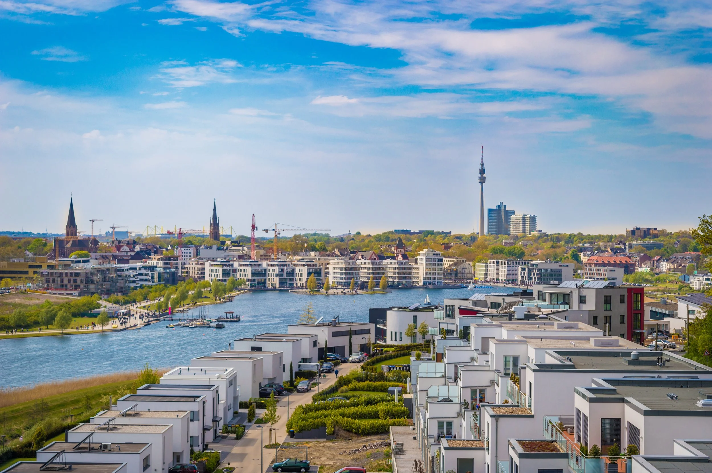 Stadtansicht mit Fluss, modernen Wohnhäusern im Vordergrund, Kirchen mit Spitzdächern, Fernsehturm und Hochhäuser im Hintergrund, bei blauem Himmel.