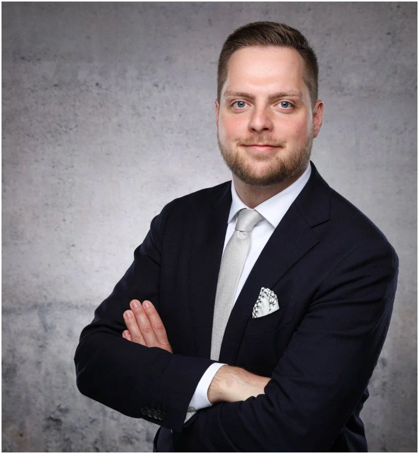 Ein Mann in formeller Kleidung mit angelegten Armen vor grauem Hintergrund.