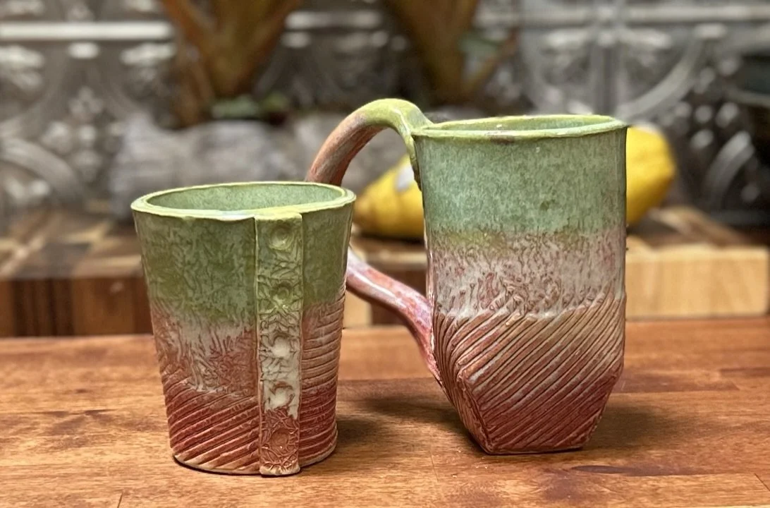 Two handmade ceramic mugs with textured design, featuring green and brown hues, displayed on a wooden surface.