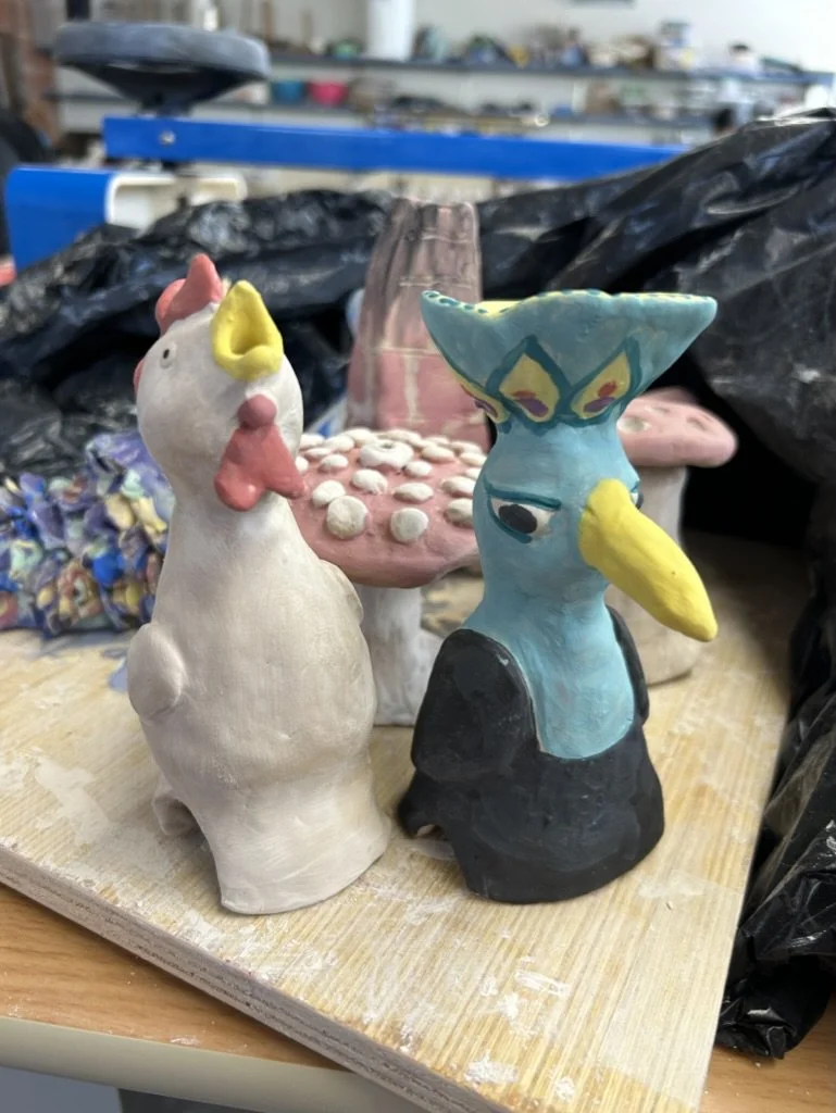 Handmade clay figurines of a chicken and a bird with a mushroom background, placed on a wooden surface in a workshop setting.