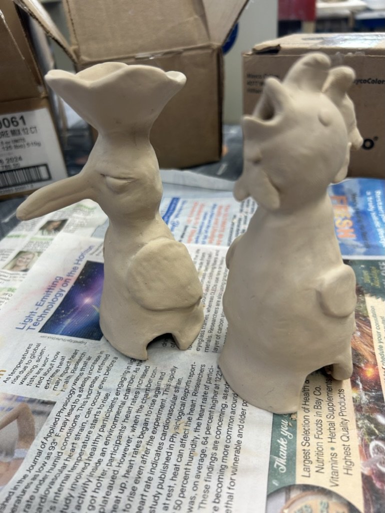 Two unfinished ceramic sculptures on newspaper, one with a long nose and the other with a tooth-shaped head.
