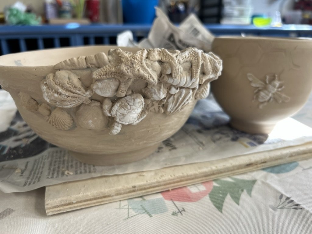 Ceramic pottery with detailed sea-themed sculptures, including starfish and shells, and a bee motif on another piece, resting on newspapers.