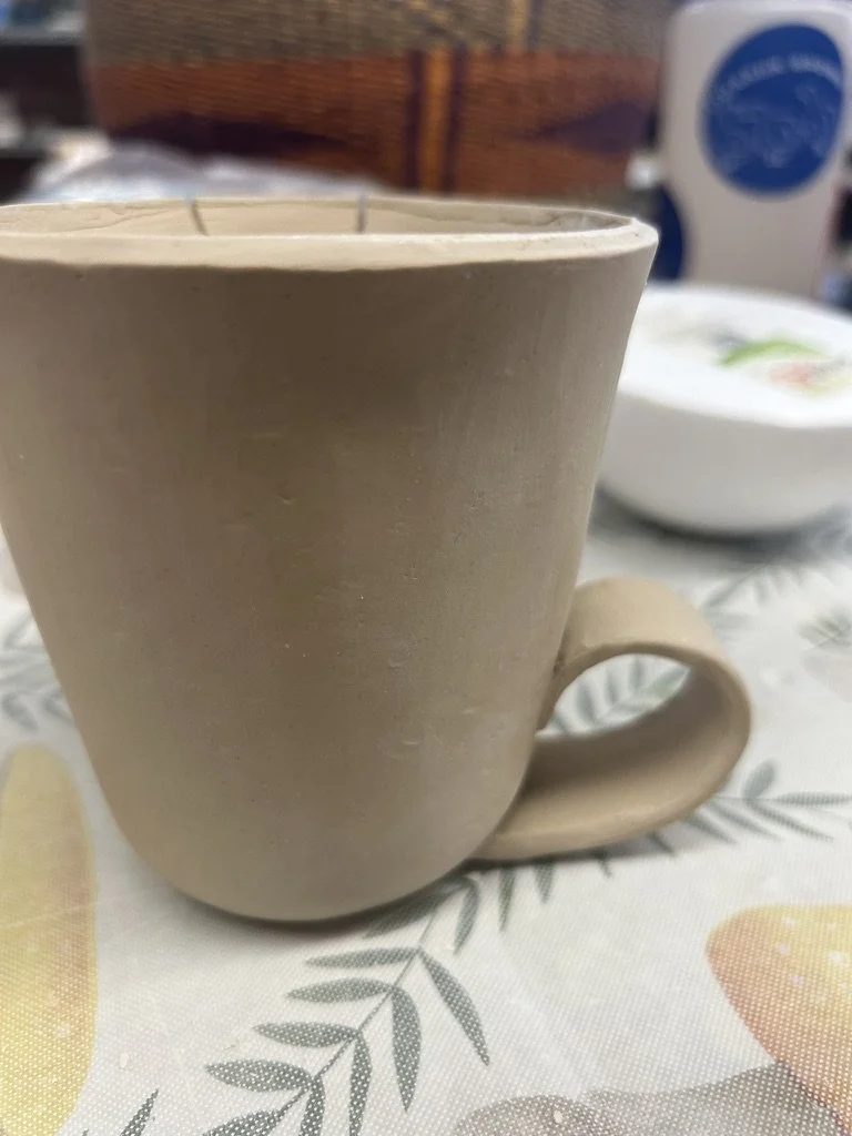 Unfinished ceramic mug with a handle on a patterned tablecloth.