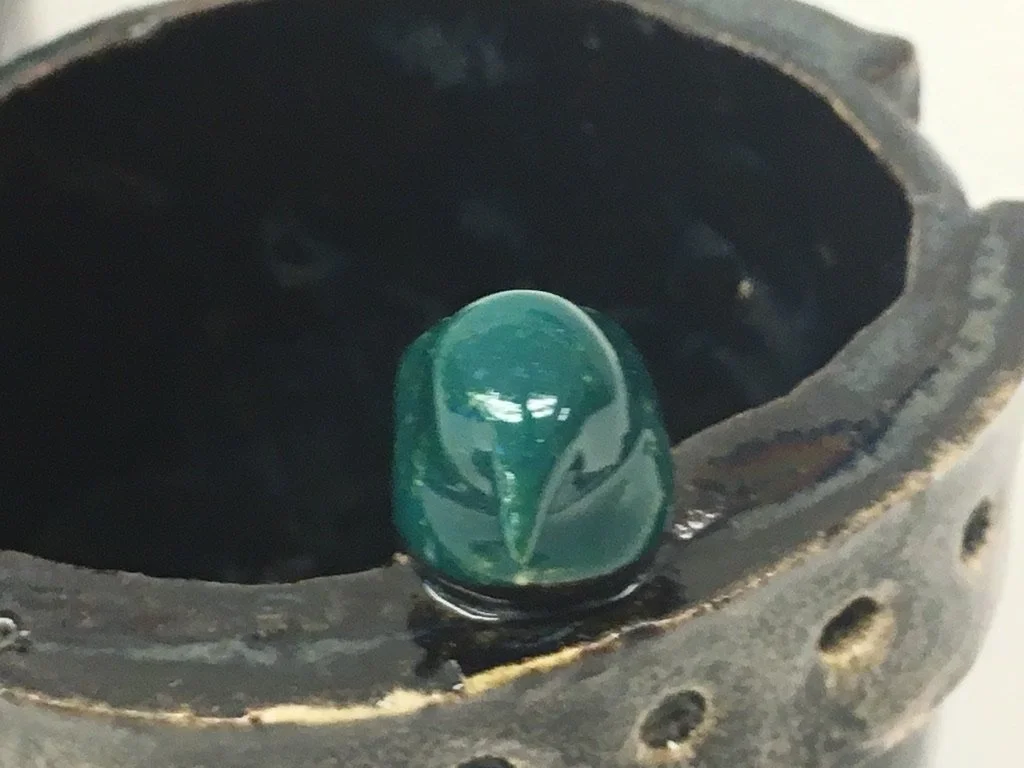 A small carved green gemstone or ornament sitting on the edge of a black, round object with a textured surface and holes.