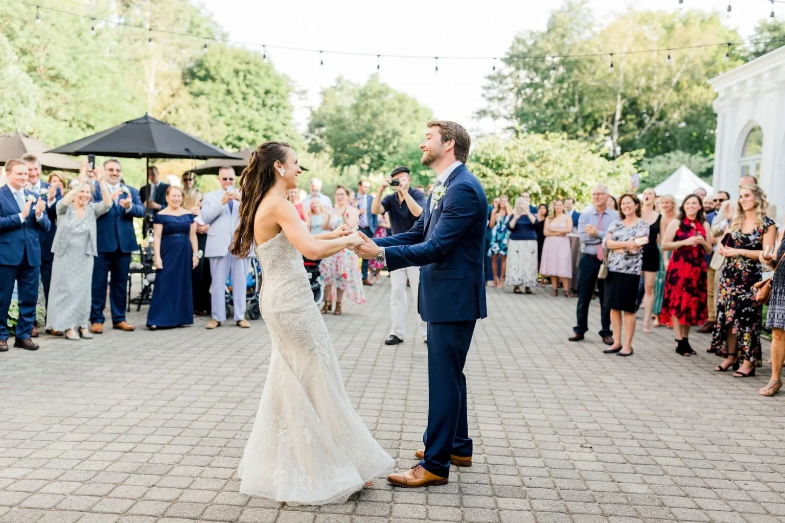 A wedding portrait featuring a couple and two children. The groom is in a gray suit with a black bow tie, and the bride is wearing a strapless white gown with floral lace and holding a bouquet of purple and pink flowers. A young girl in a white dress with floral headpiece and a boy in a white shirt with suspenders and a bow tie stand between them, all smiling and posed in front of a building entrance.