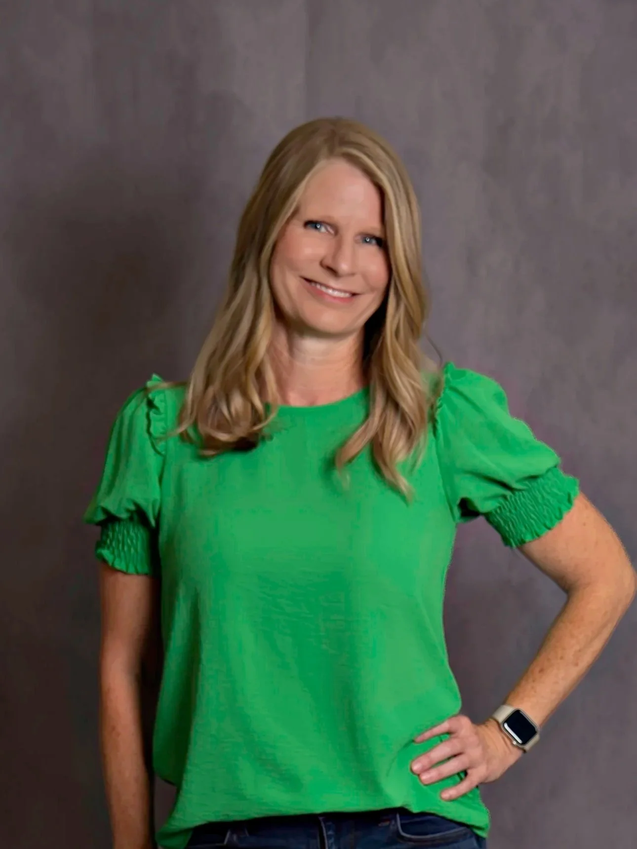 Smiling person with long blonde hair wearing a green top, standing outdoors with a blurred green background.