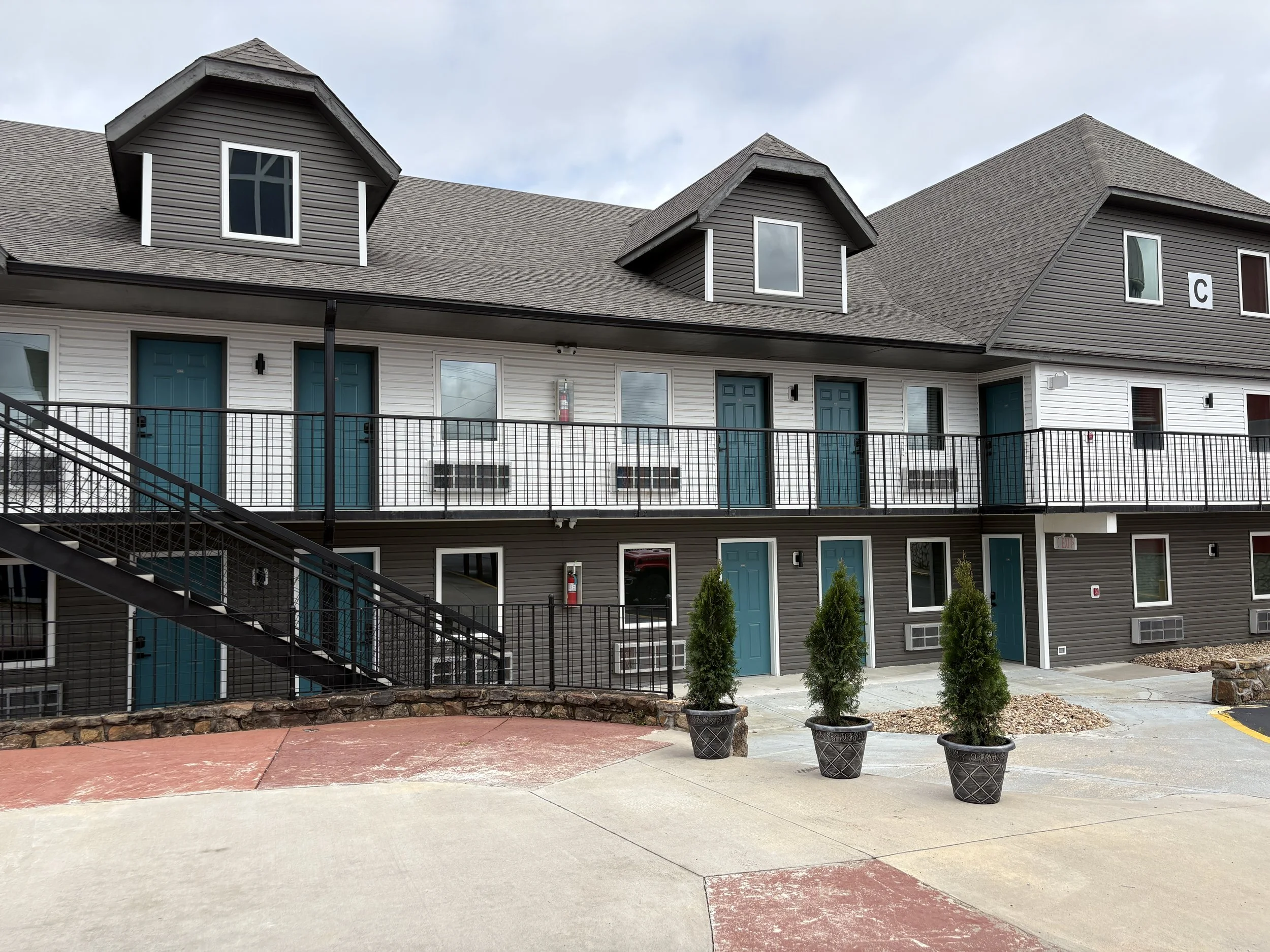 Exterior of a multiple-story apartment building with gray siding, teal doors, and several windows. The building features a black metal staircase and railing. In front, there are three potted evergreen trees placed on a concrete sidewalk.