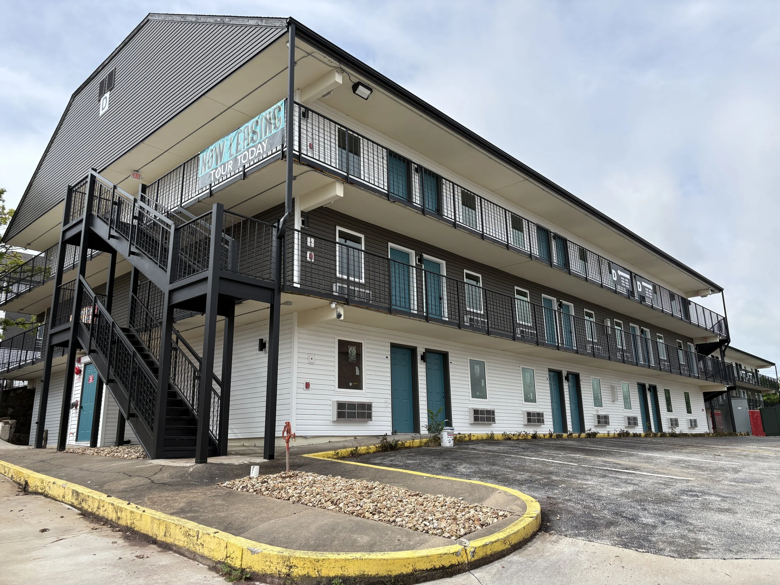A multi-story apartment building with black exterior siding and blue doors, featuring outdoor metal staircases and railings. There is a sign on the balcony that reads "NOW LEASING TOUR TODAY." The parking lot in front has a yellow curb and some small