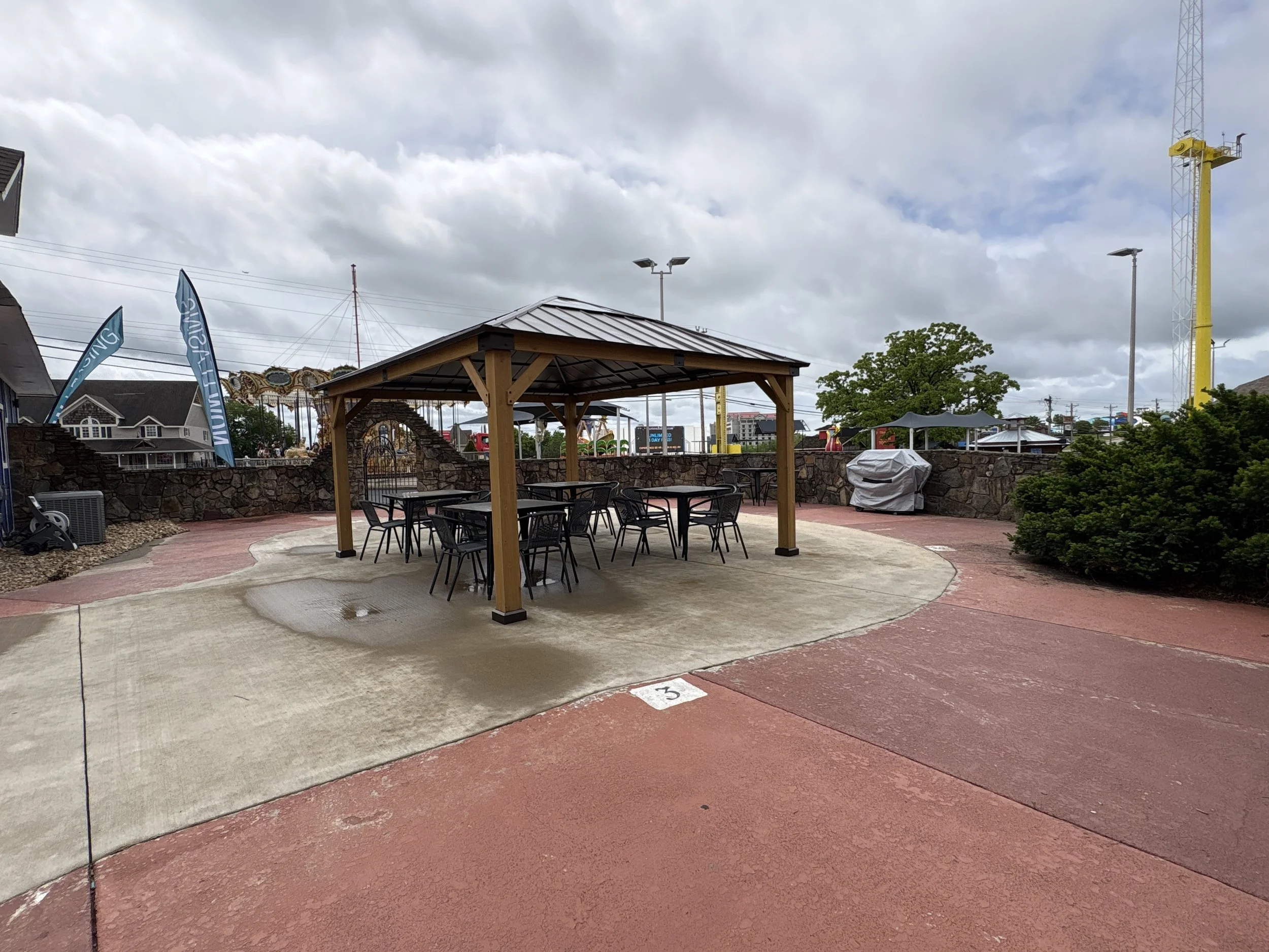 Photo of an outdoor amusement park area on a cloudy day, including a covered seating area with tables and chairs, roller coaster tracks in the background, and flags and structures related to the amusement park.