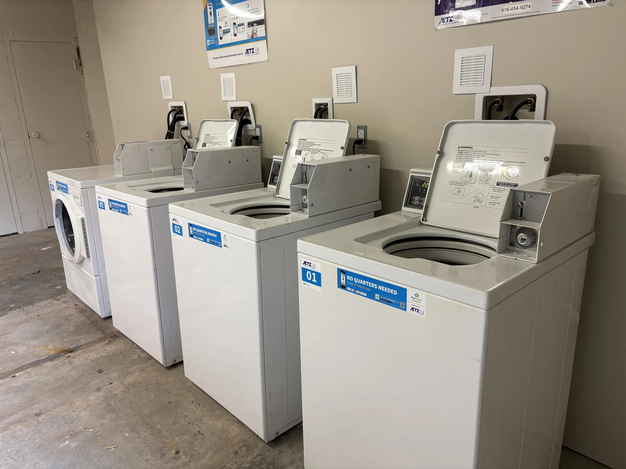 Four white commercial washing machines lined up against a beige wall in a laundry facility, with instructions and signage above each machine.