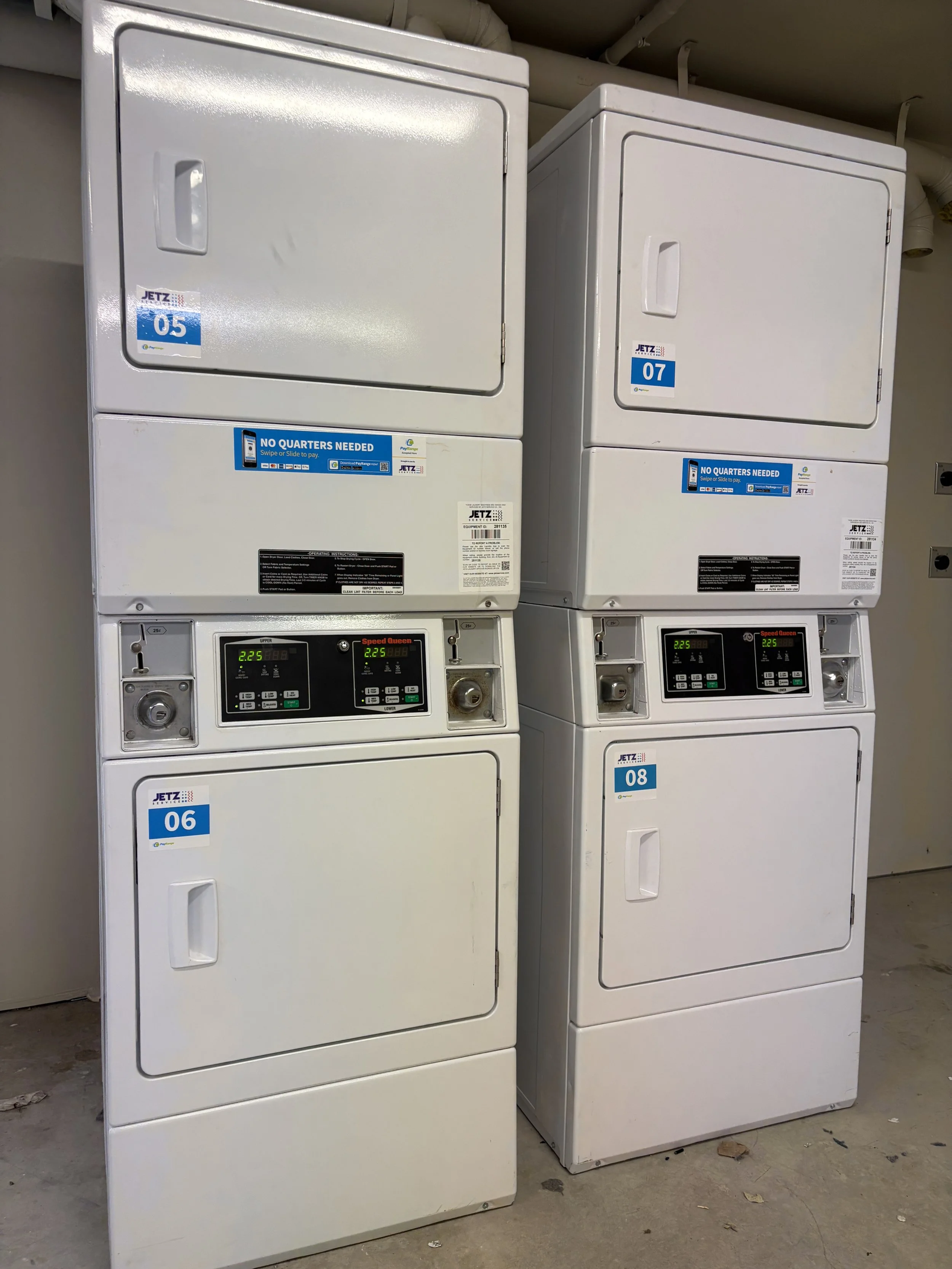 Two white commercial dryers stacked in a laundry room with control panels and coin slots, labeled with numbers 06 and 08.
