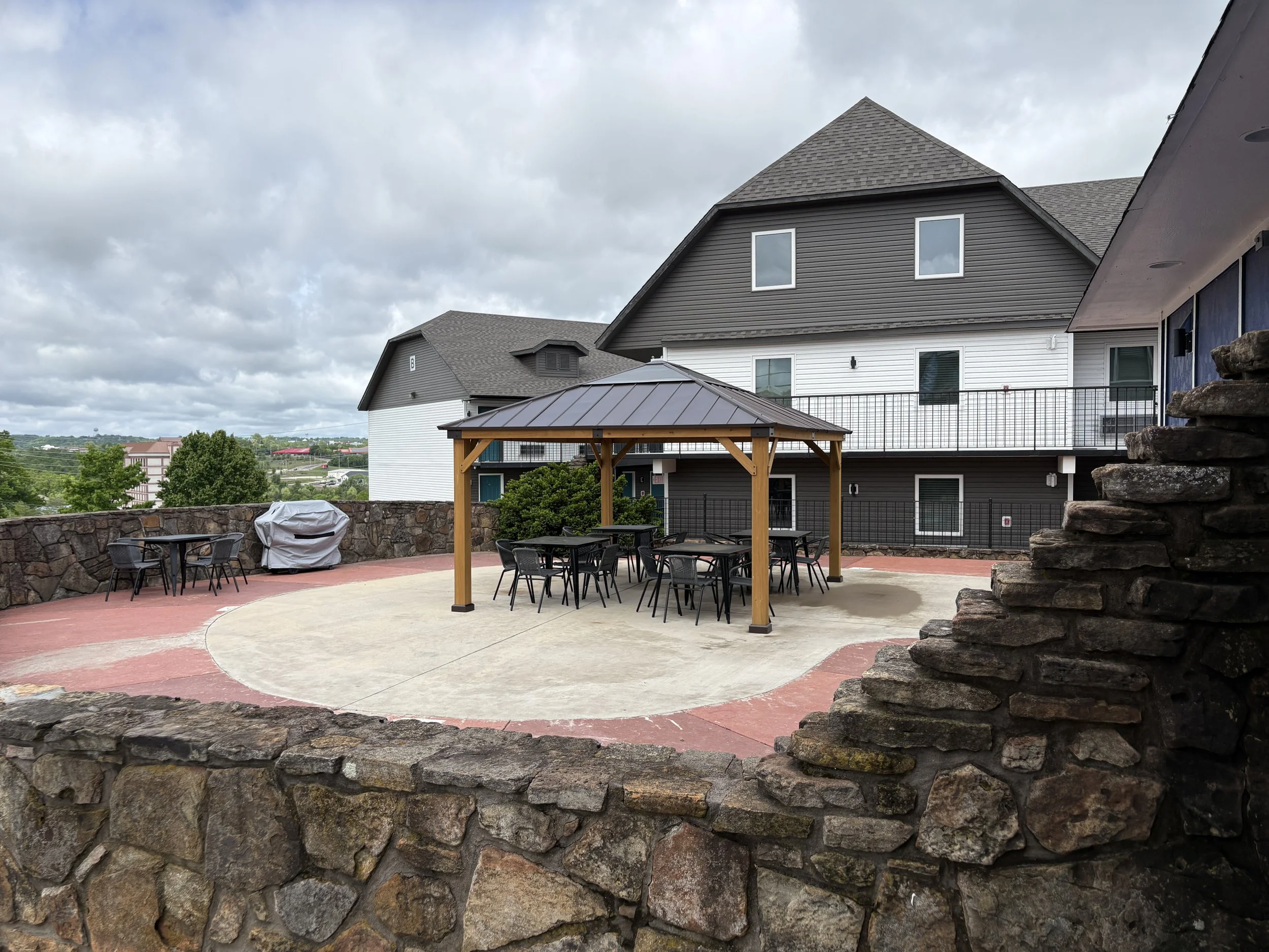 Outdoor patio area with a stone wall, a table with black chairs, a covered structure, and residential buildings in the background under cloudy skies.