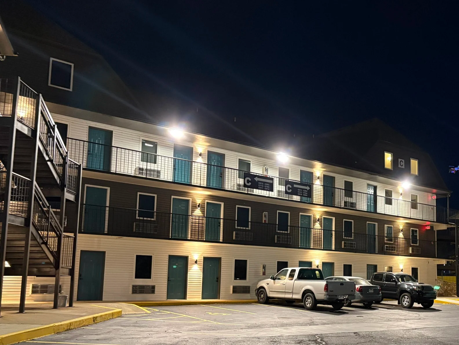 Night view of a three-story motel with parked cars in the parking lot, external corridor, doors, and illuminated light fixtures on the building.