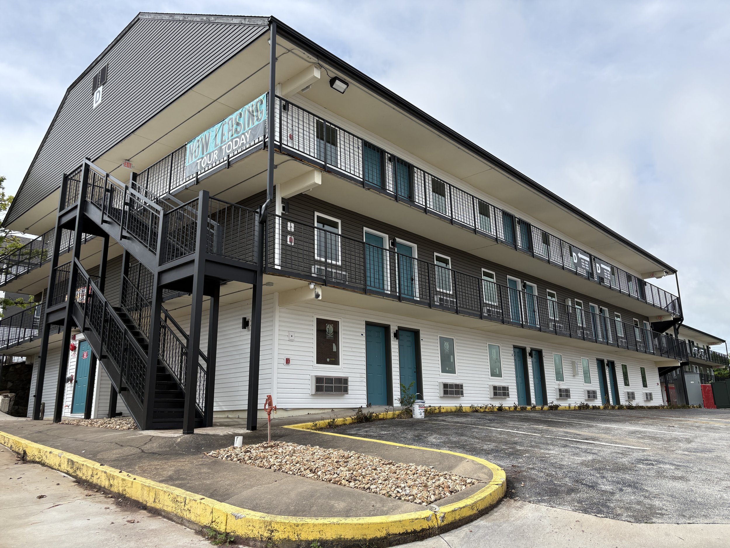Multi-story building with black metal railings, exterior stairs, blue doors, and signs promoting a tour now leasing. The building has a modern design with gray and white siding, and the parking lot in front has a yellow curb and some gravel areas.