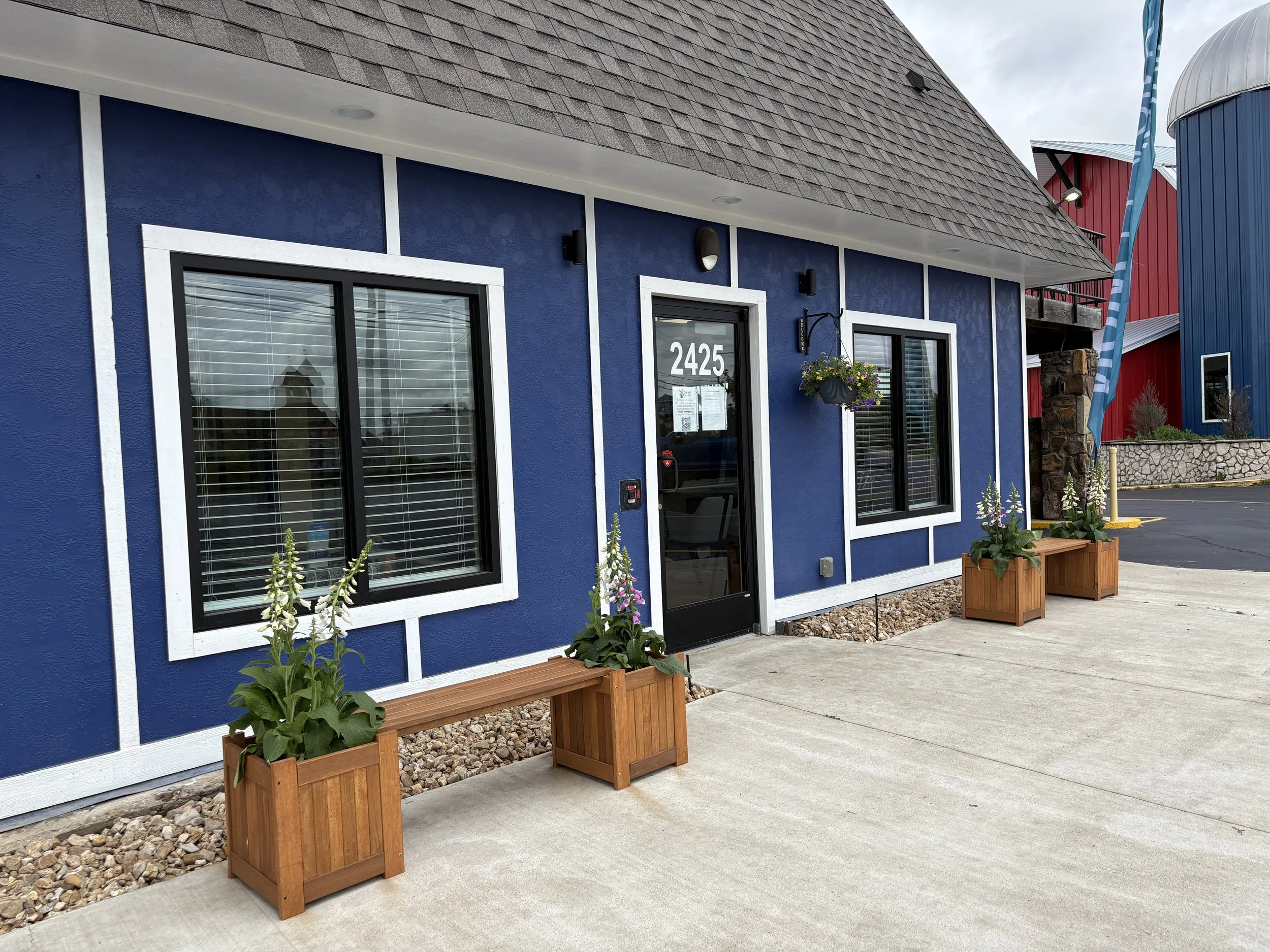 Front of a blue building with white trim, two windows, a door with the number 2425, and flower boxes with plants and flowers. There are wooden benches outside the building, and the sidewalk and parking lot are visible.