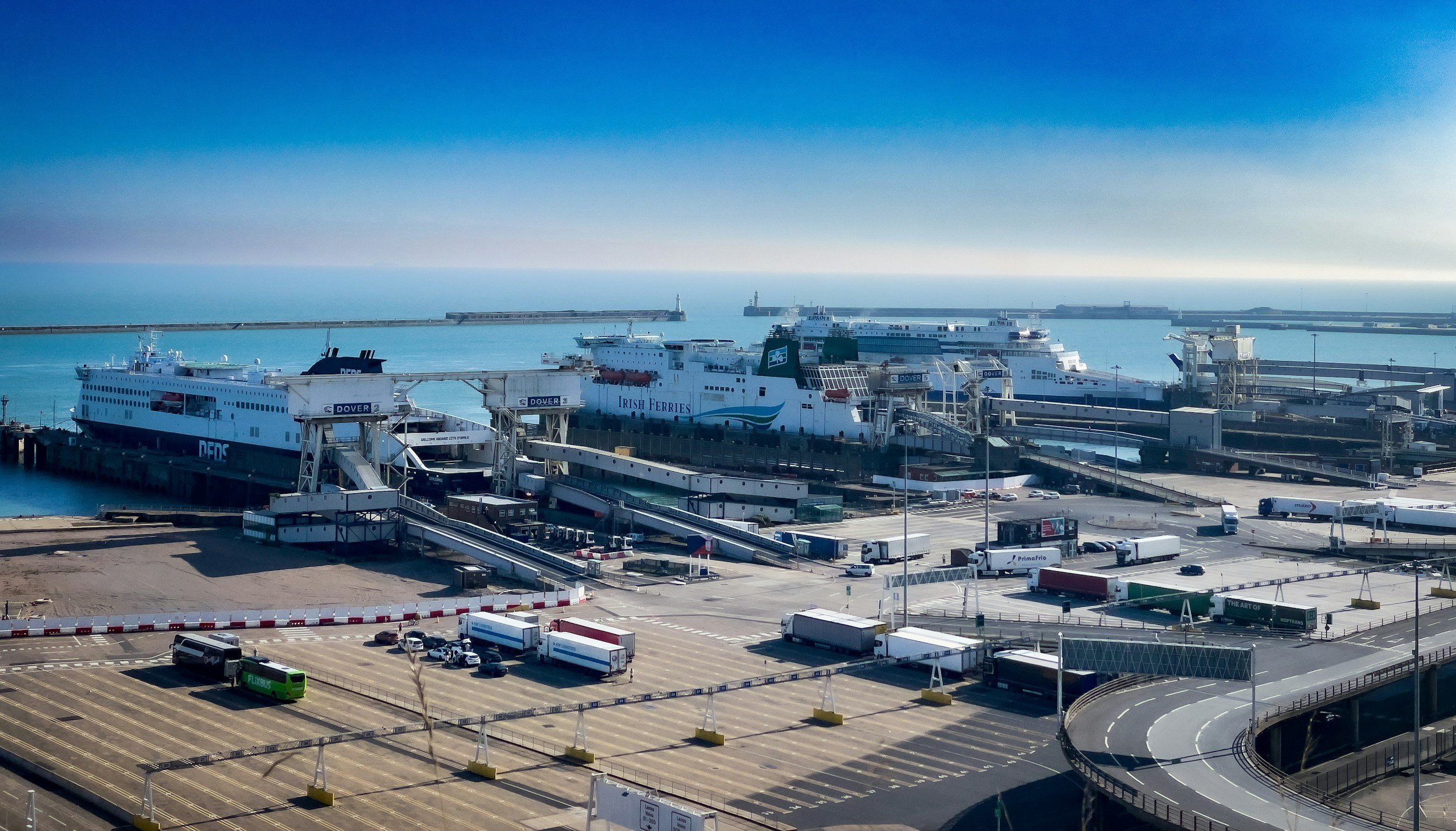 View of a ferry port with multiple ferries, including Irish Ferries and DFDS, docked at the pier, with vehicles and passenger buses in the parking area. The sea and harbor infrastructure are visible in the background under a clear blue sky.