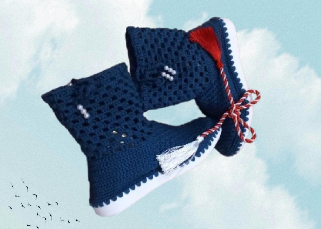 A pair of crocheted navy blue baby booties with white decorative beads, red and white twine, and a white sole, floating against a partly cloudy sky with a flock of birds in the distance.