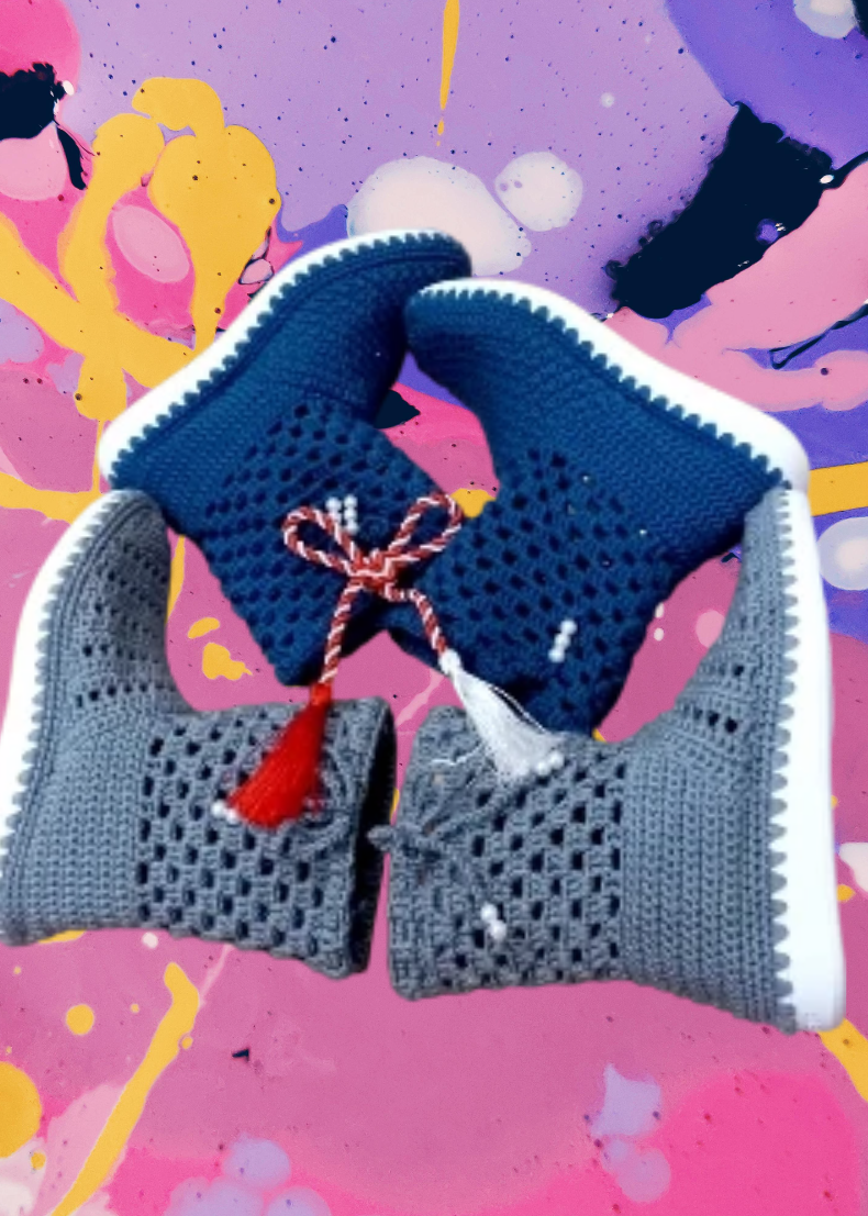 Four small, knitted baby shoes, two in dark blue and two in gray, accented with red and white tassels, placed on a colorful, abstract painted background.