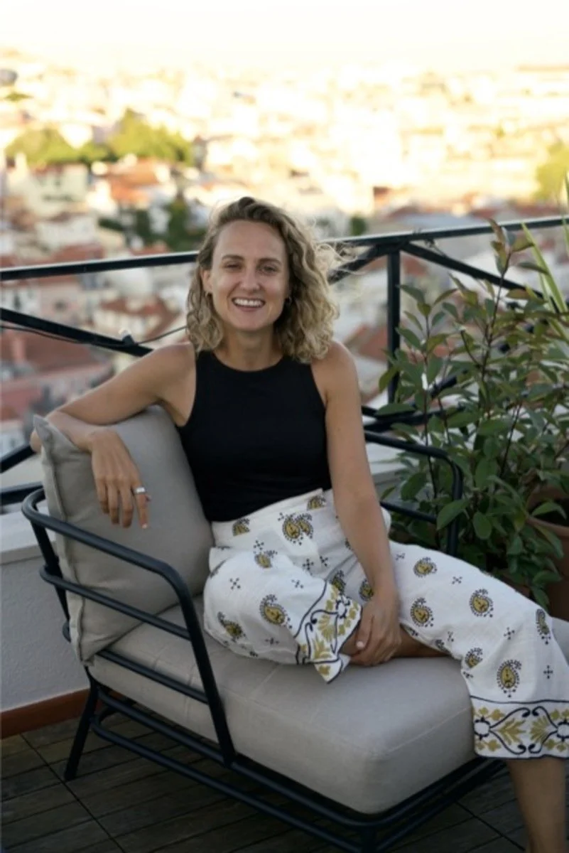 Woman sitting on an outdoor balcony sofa with a cityscape in the background, smiling at the camera.