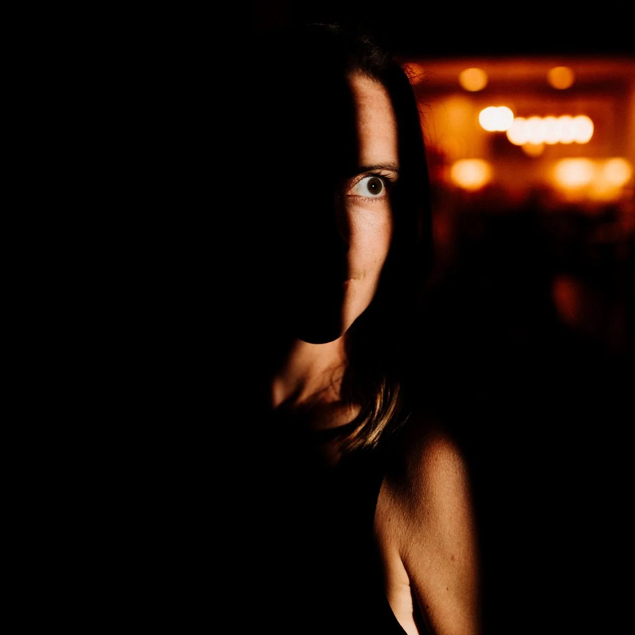 A woman with dark hair partially hidden in shadows, with one eye visible and illuminated by warm ambient lighting in the background.