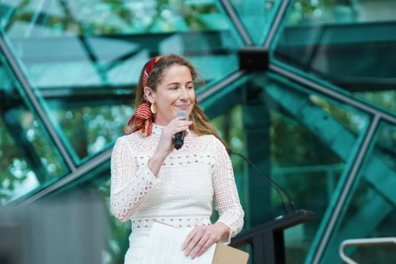 A woman with long, wavy brown hair held back with a red and white polka dot headband, speaking into a microphone at a lectern outdoors with modern glass architecture and green trees in the background.
