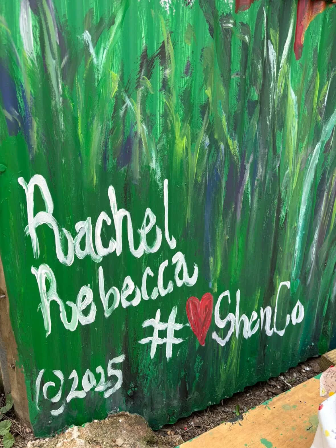 A green painted sign with white and red handwritten text that reads, 'Rachel! Rebecca #Shenco' with a red heart symbol, outdoors with some dirt and wood at the base.