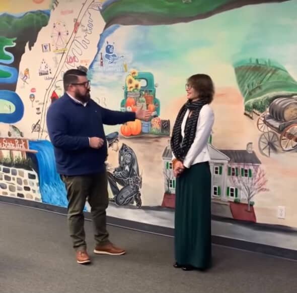 Two people standing in front of a colorful mural featuring landscapes, a waterfall, and various local landmarks. The man is gesturing while speaking to the woman.