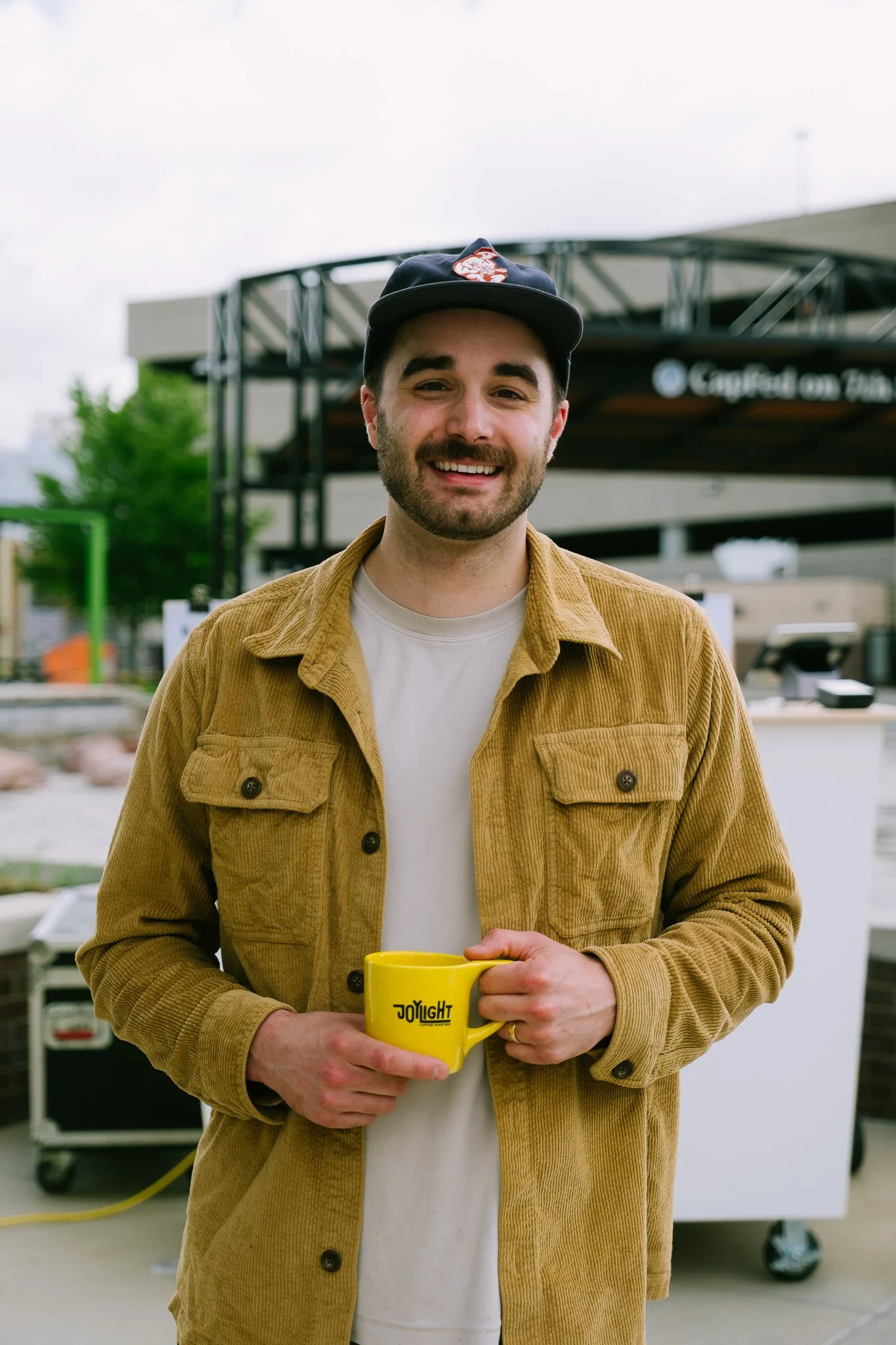 A smiling young man with a beard wearing a dark baseball cap and a beige jacket, holding a yellow coffee mug with black text, outdoors at a coffee stand.