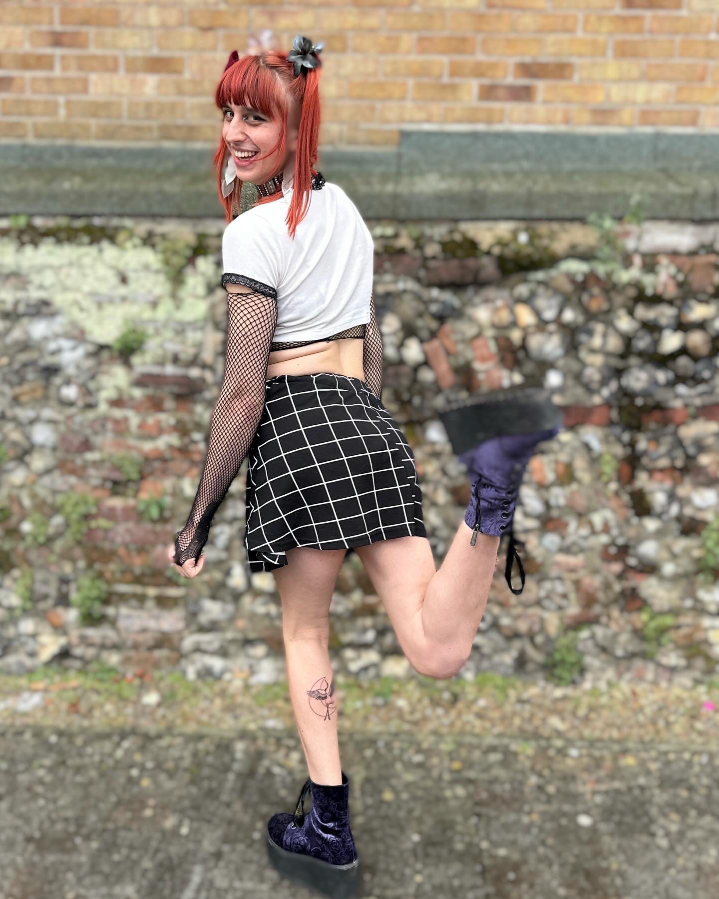 Person with red hair in a playful pose, wearing a white top, black checkered skirt, and platform boots, standing against a brick wall.