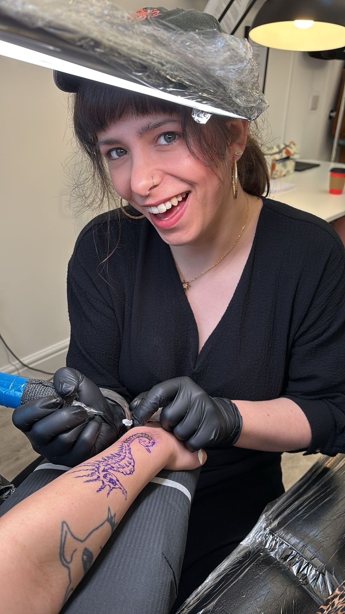 Tattoo artist wearing black gloves, smiling, while working on a tattoo of a scorpion on a person's arm in a tattoo studio.