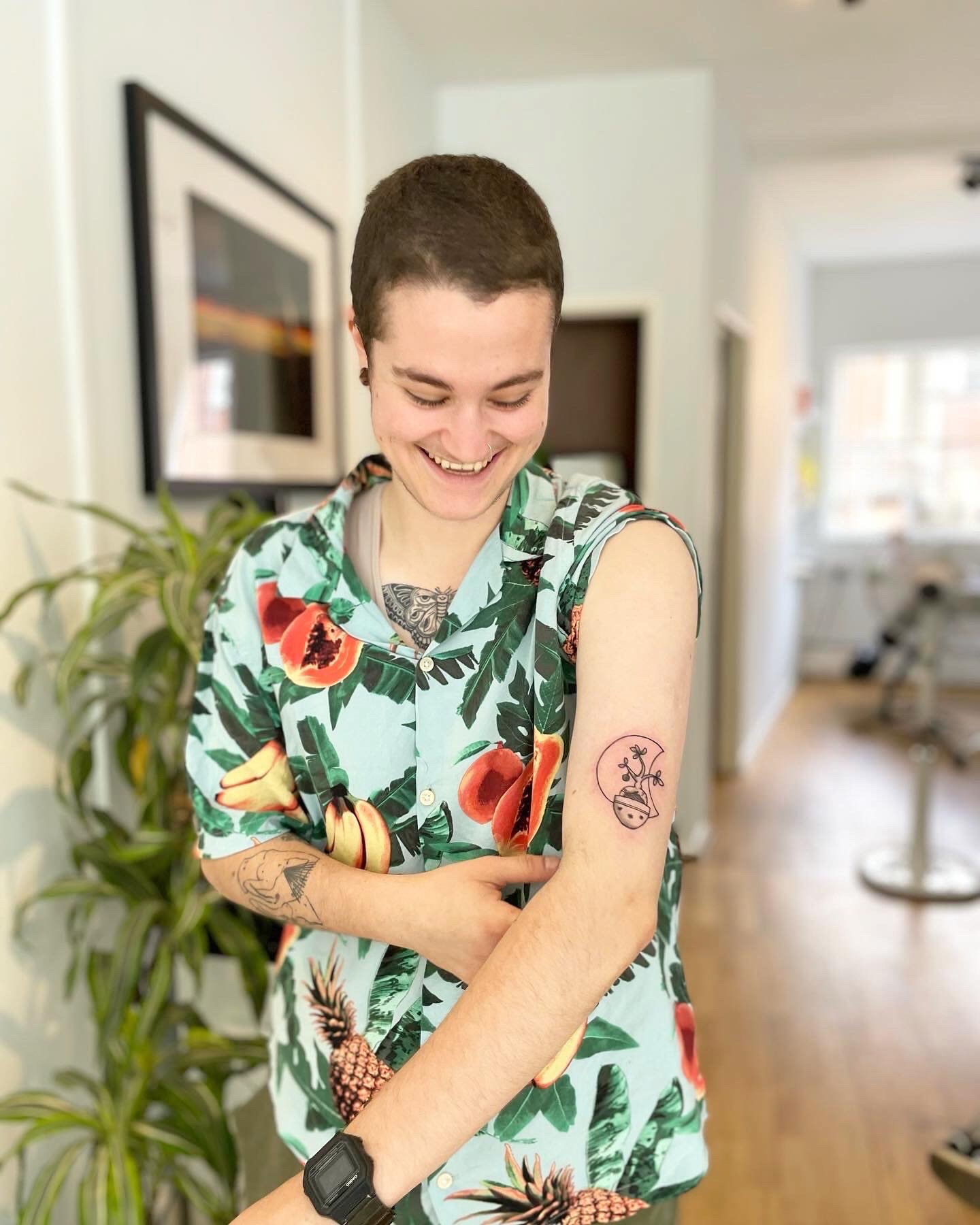 Person smiling, looking at tattoo on upper arm, wearing a colorful patterned shirt with fruit design, standing in a bright room with a plant visible in the background.